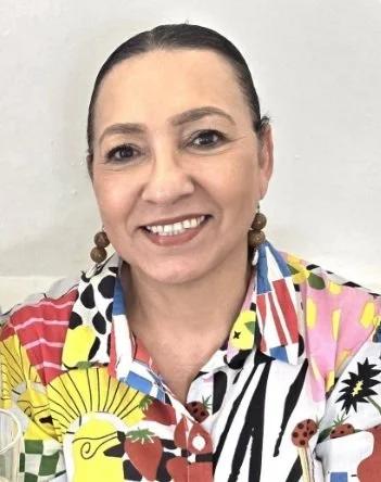 A woman with dark hair pulled back, smiling, wearing a colorful, patterned shirt with various bright colors and designs.