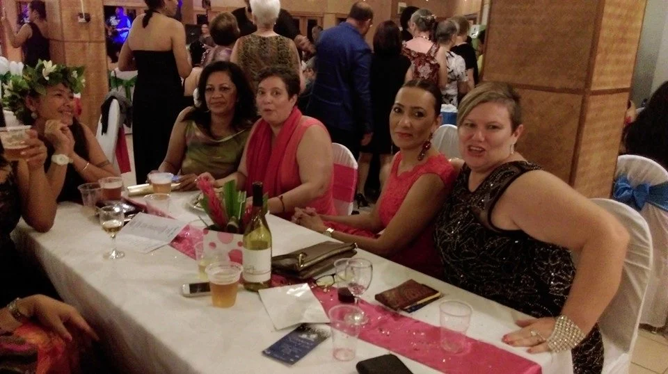 Five women seated at a banquet table during a celebration or party, with guests standing in the background, decorated with flowers and pink accents.