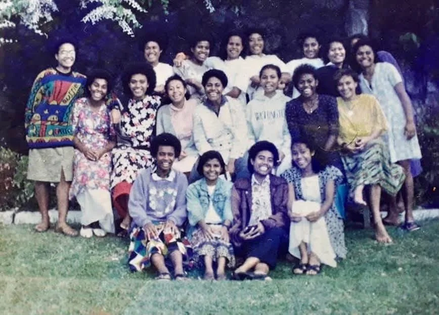 Group of 16 people gathered outdoors on a grassy area, smiling, with trees in the background.