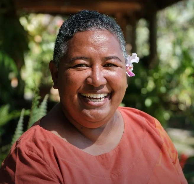 Smiling woman with short curly hair, dark skin, wearing a pink flower behind her ear and a coral-colored shirt, outdoors in a lush green garden.
