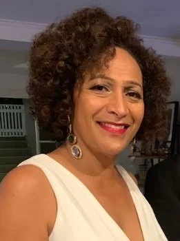 A smiling woman with curly hair wearing a white sleeveless top and dangling earrings, standing indoors.