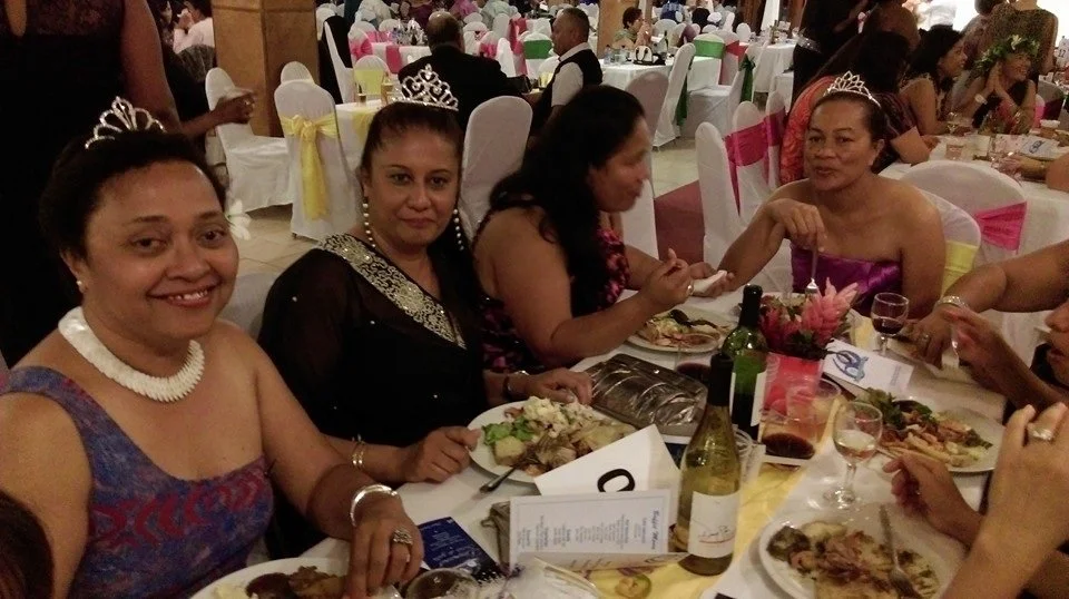 Four women wearing tiaras and formal dresses seated at a decorated banquet table during a celebration or wedding reception.