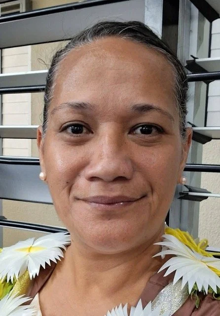 Close-up of a woman with pearl earrings, wearing a dress with white and yellow flower decorations on the shoulders, standing outdoors near a gray building with window shutters.