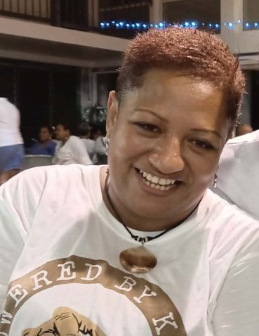 Close-up of a smiling woman wearing a white T-shirt with a circular logo, and a small brown stone necklace. She is indoors with a blurred background of other people.