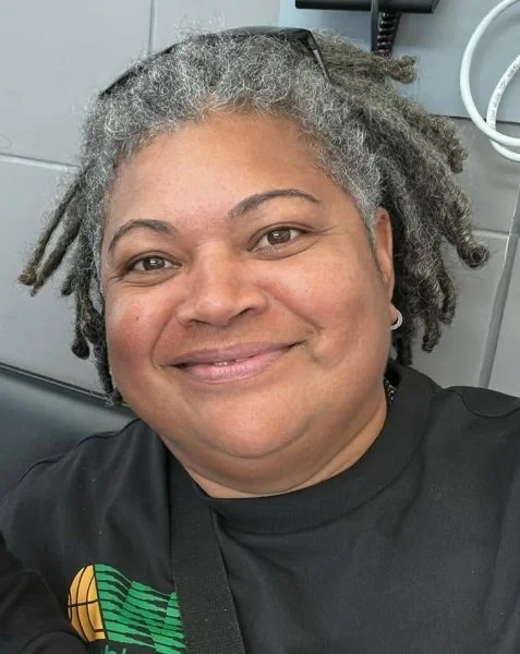 Close-up photo of an smiling woman with gray dreadlocks, wearing a black shirt, inside a room with gray walls and medical or technical equipment.