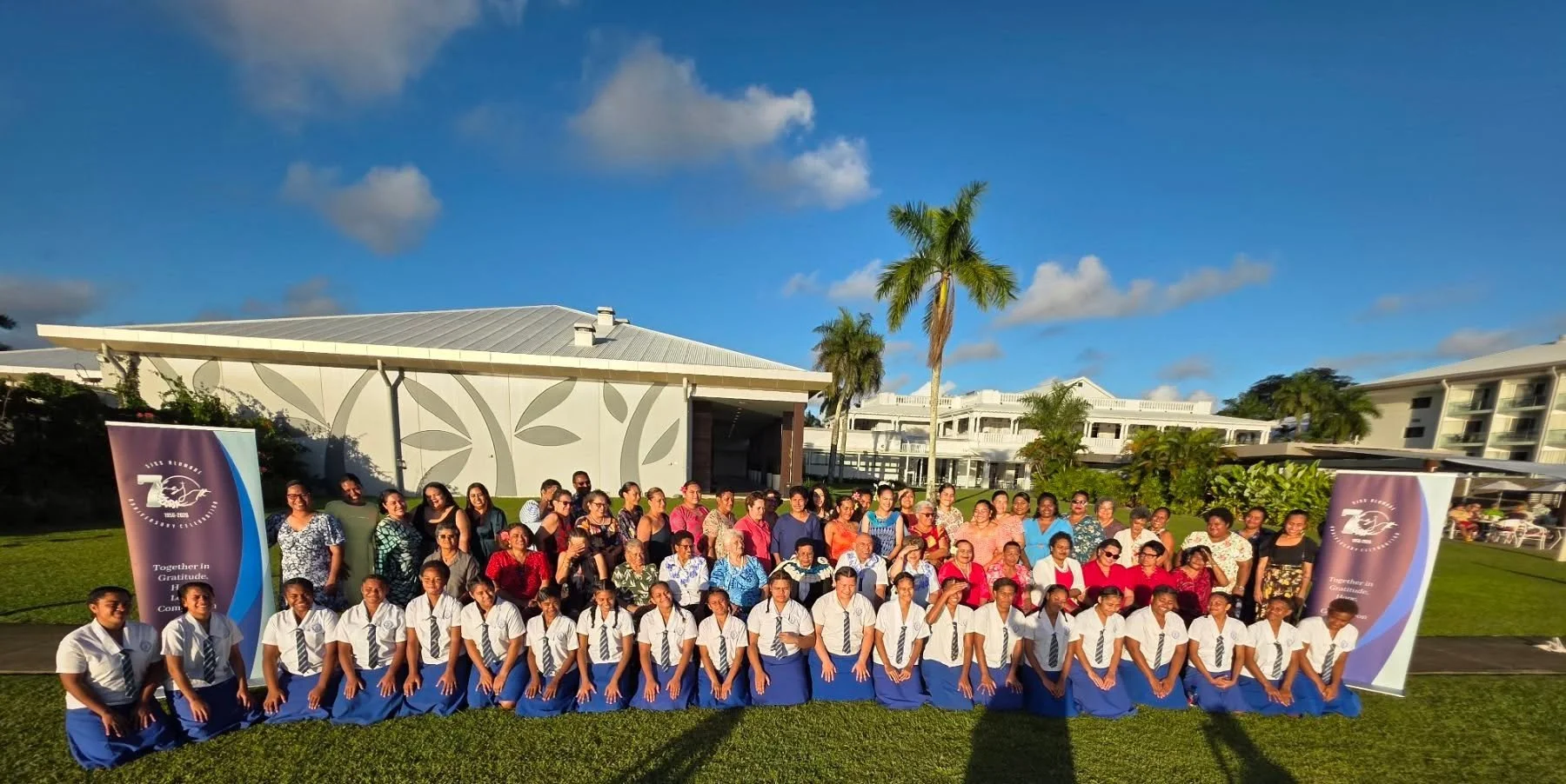Group photo of students and teachers outdoors on green grass, with two purple banners in the foreground, large white building, palm trees, and partly cloudy blue sky in the background.