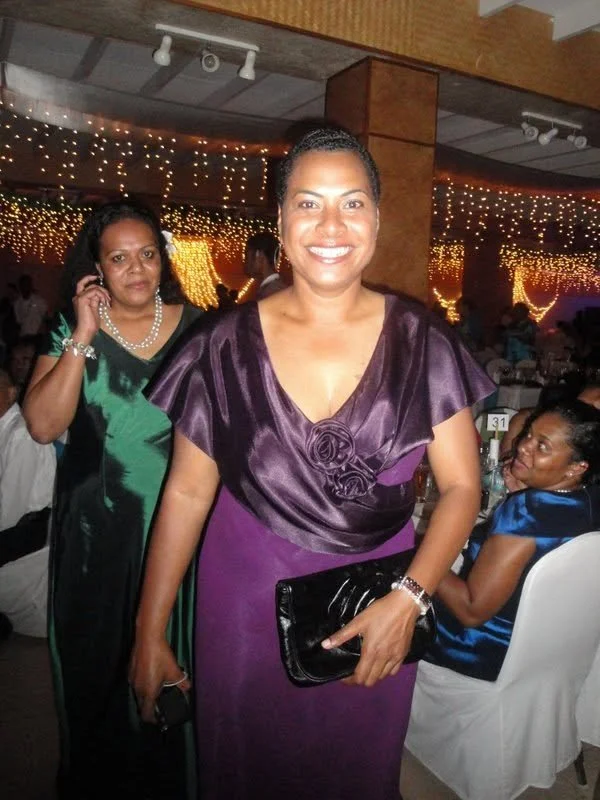Woman in a purple dress smiling at a social gathering or celebration, with other guests and festive lighting in the background.
