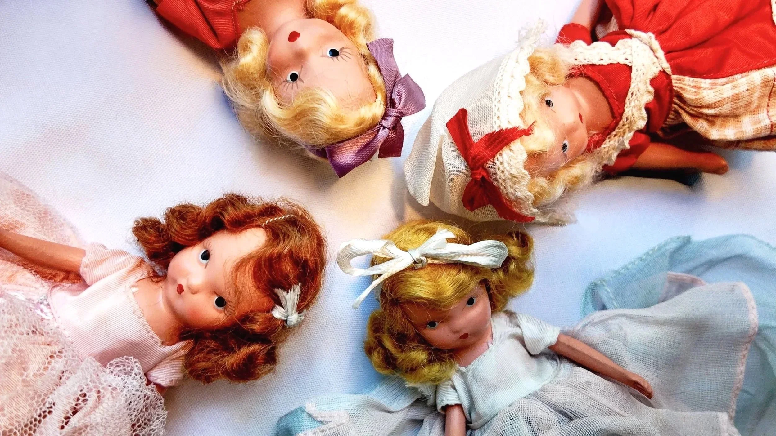 Four vintage porcelain dolls with curled hair and traditional clothing arranged on white fabric.