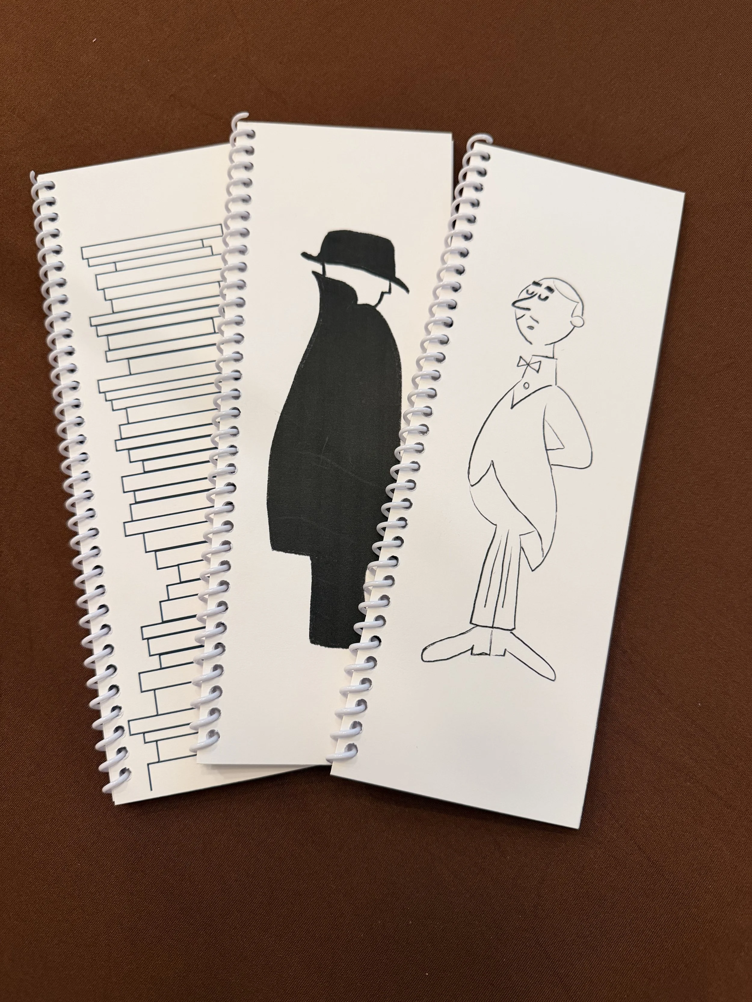 Three notebooks with sketches on a brown surface. The first has a geometric pattern, the second features a black silhouette of a woman with a wide-brimmed hat, and the third shows a line drawing of a man with a bow tie in a confident pose.