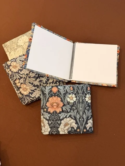 A stack of four patterned notebooks with floral designs, one open with blank pages, placed on a wooden surface.