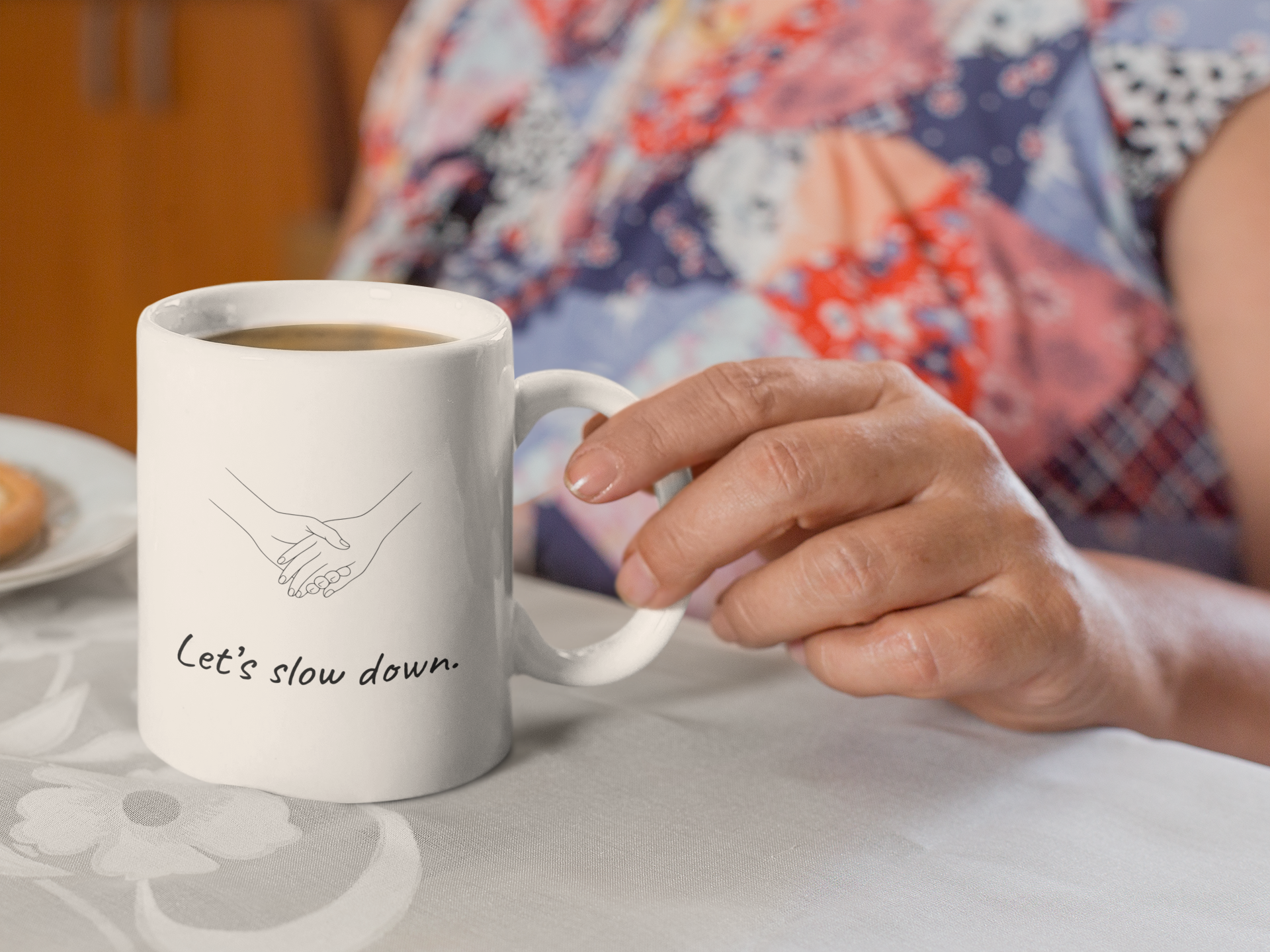 Close-up of a person holding a white coffee mug with a black line drawing of two hands holding, and text that says 'Let's slow down.' The person has a light skin tone and is wearing a colorful, patterned shirt. There is a plate with food in the background, blurred.