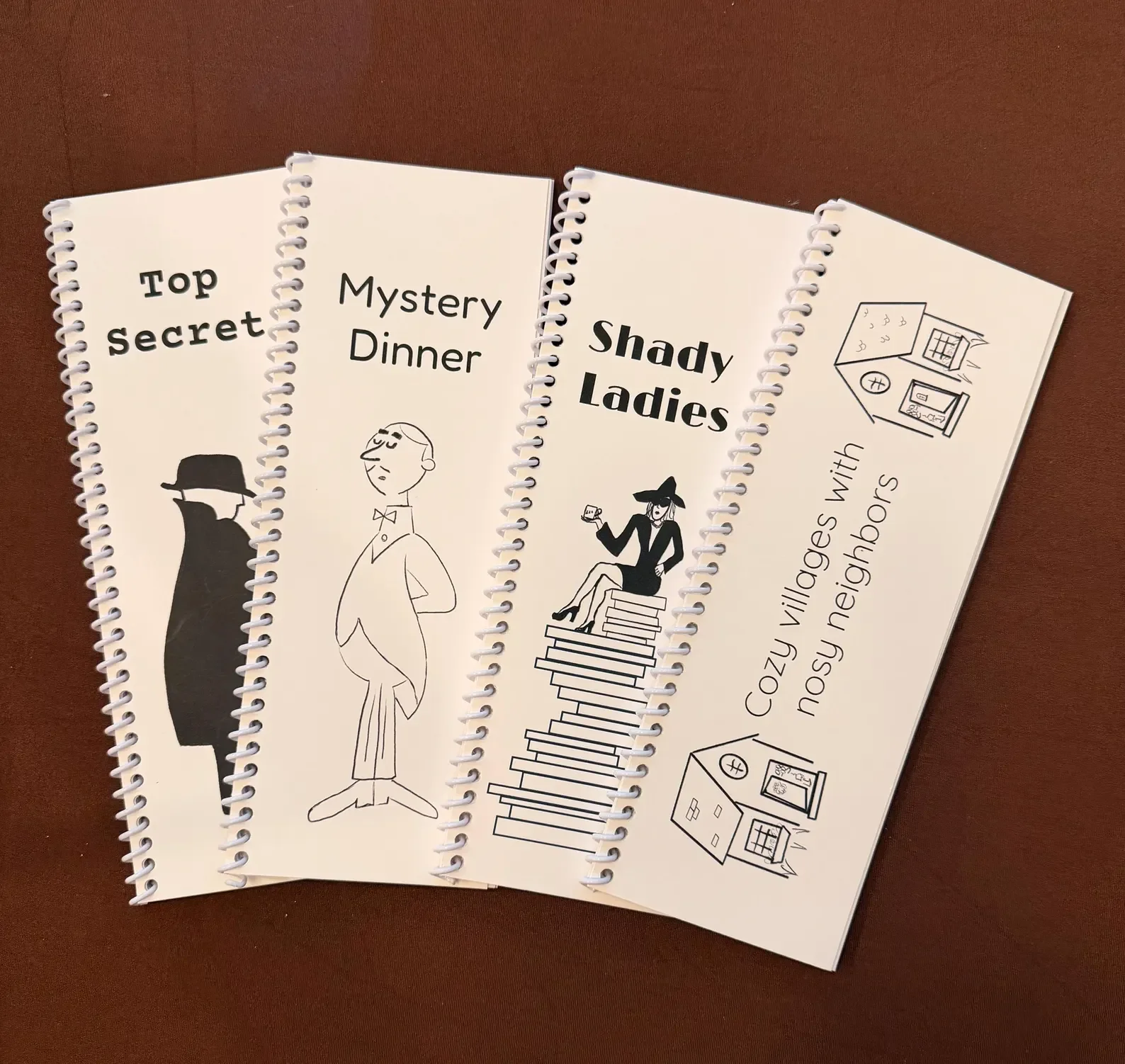 Four spiral notebooks with humorous titles and illustrations on a brown surface.