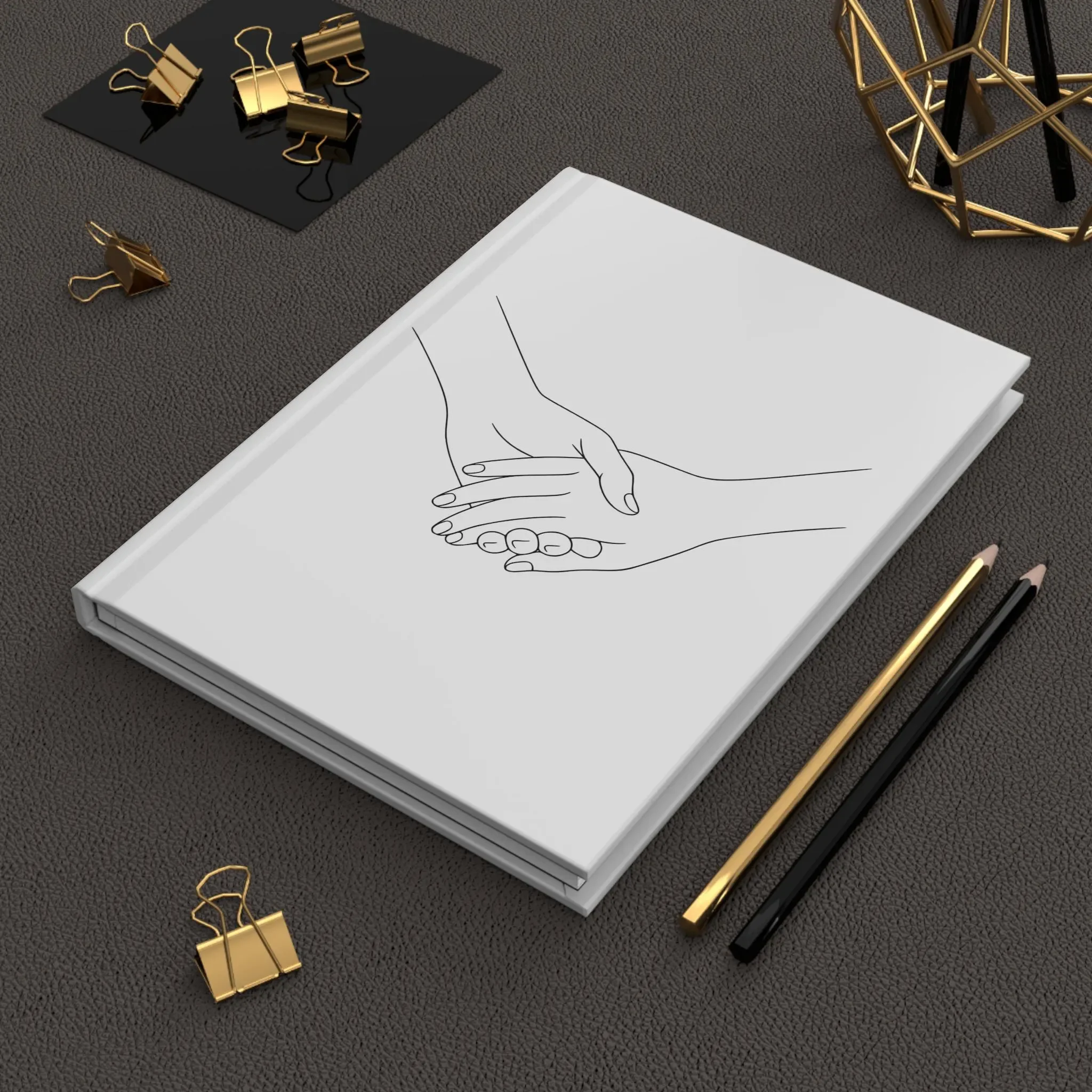 A desk with gold binder clips, a gold and black wireframe geometric sculpture, and a white sketchpad with a drawing of two hands shaking.