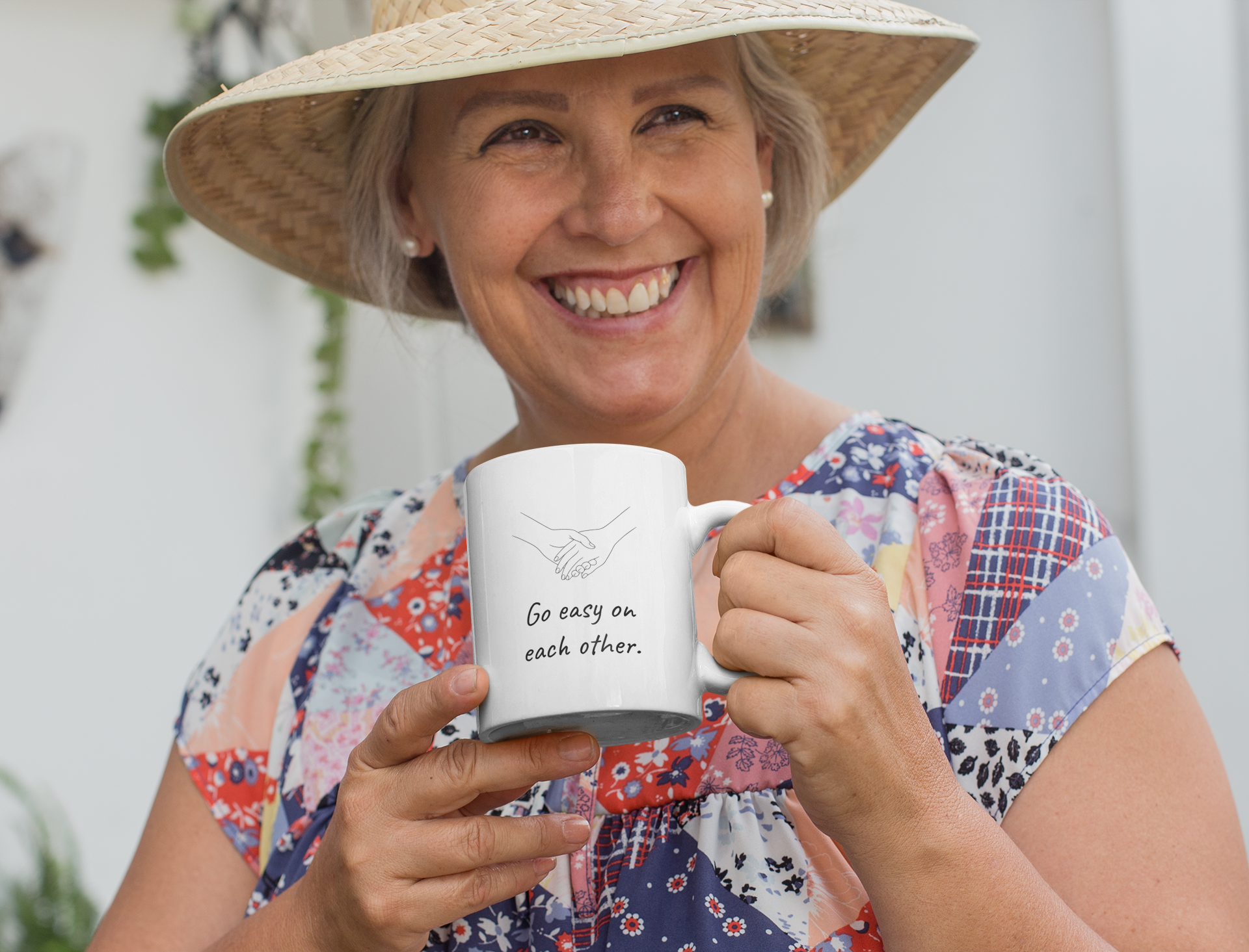 A smiling elderly woman wearing a colorful patchwork dress and a large straw hat, holding a white mug with a drawing of two hands shaking and the text 'Go easy on each other.'