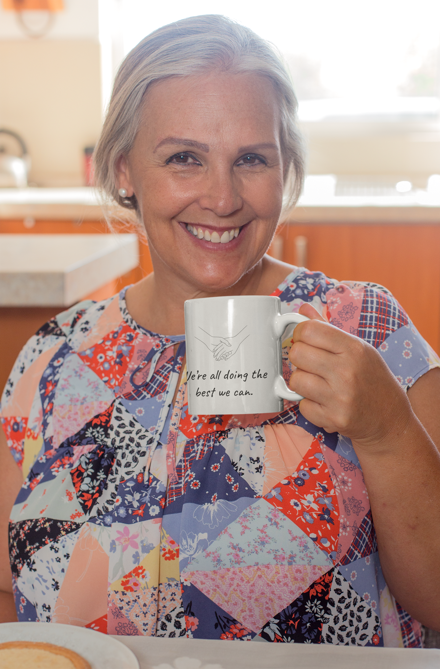 A smiling woman with short gray hair holding a white mug that has a drawing of two hands shaking and a quote on it, in a brightly lit kitchen environment.