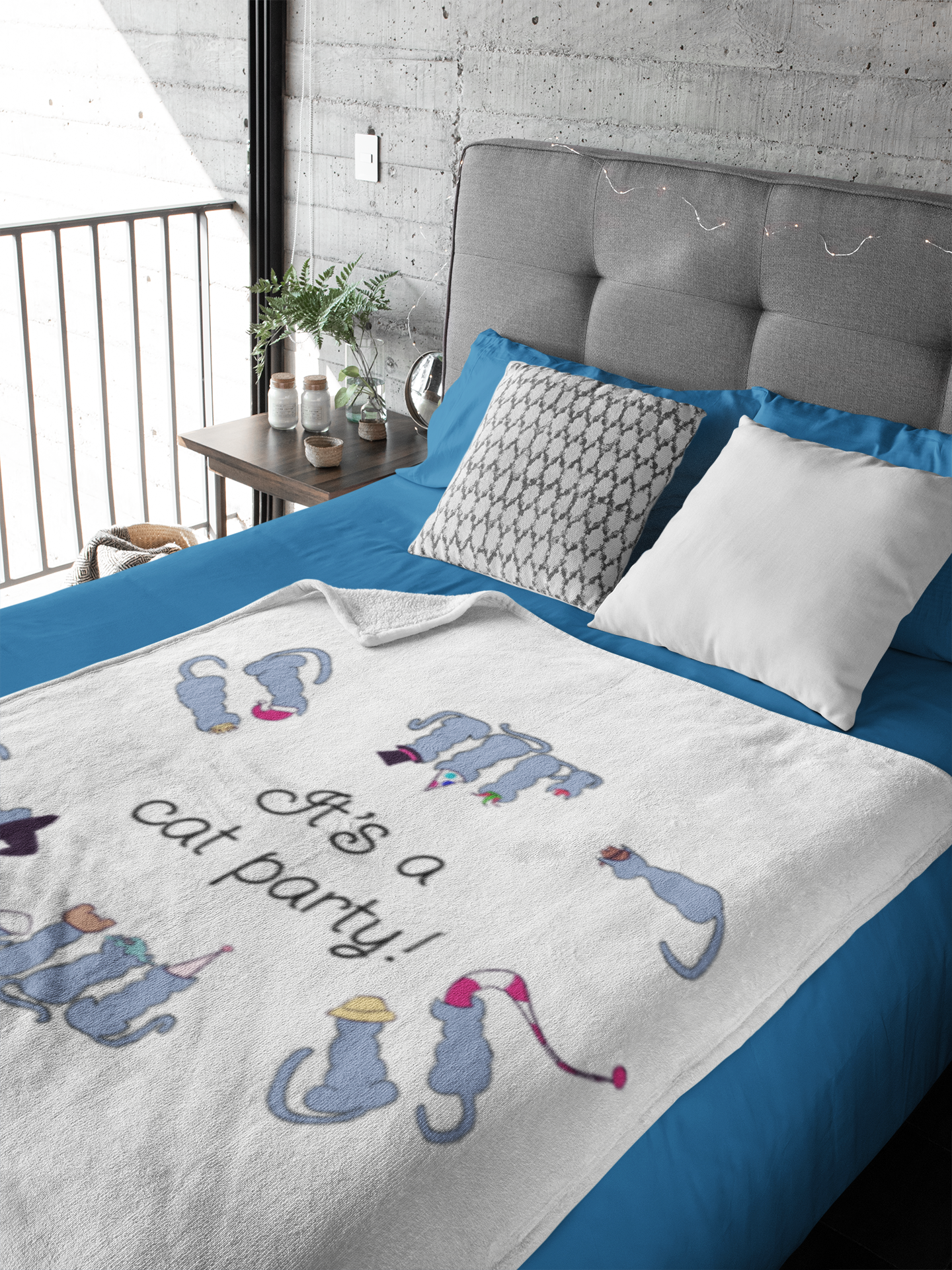 A bed with a gray upholstered headboard and blue bedding, next to a wooden nightstand with a plant, candles, and decorative items. The bed has pillows, including one with a black and white geometric pattern. A blanket with the text "He's a cat person!" and colorful animals is spread across the bed. The room features a concrete wall and a balcony with black railing.