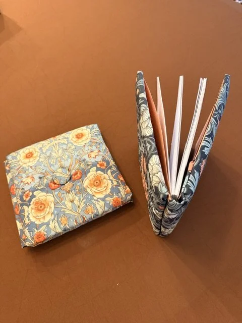 A closed floral-patterned notebook or journal on a wooden surface, next to a standing file organizer with more floral-patterned folders and some loose papers.