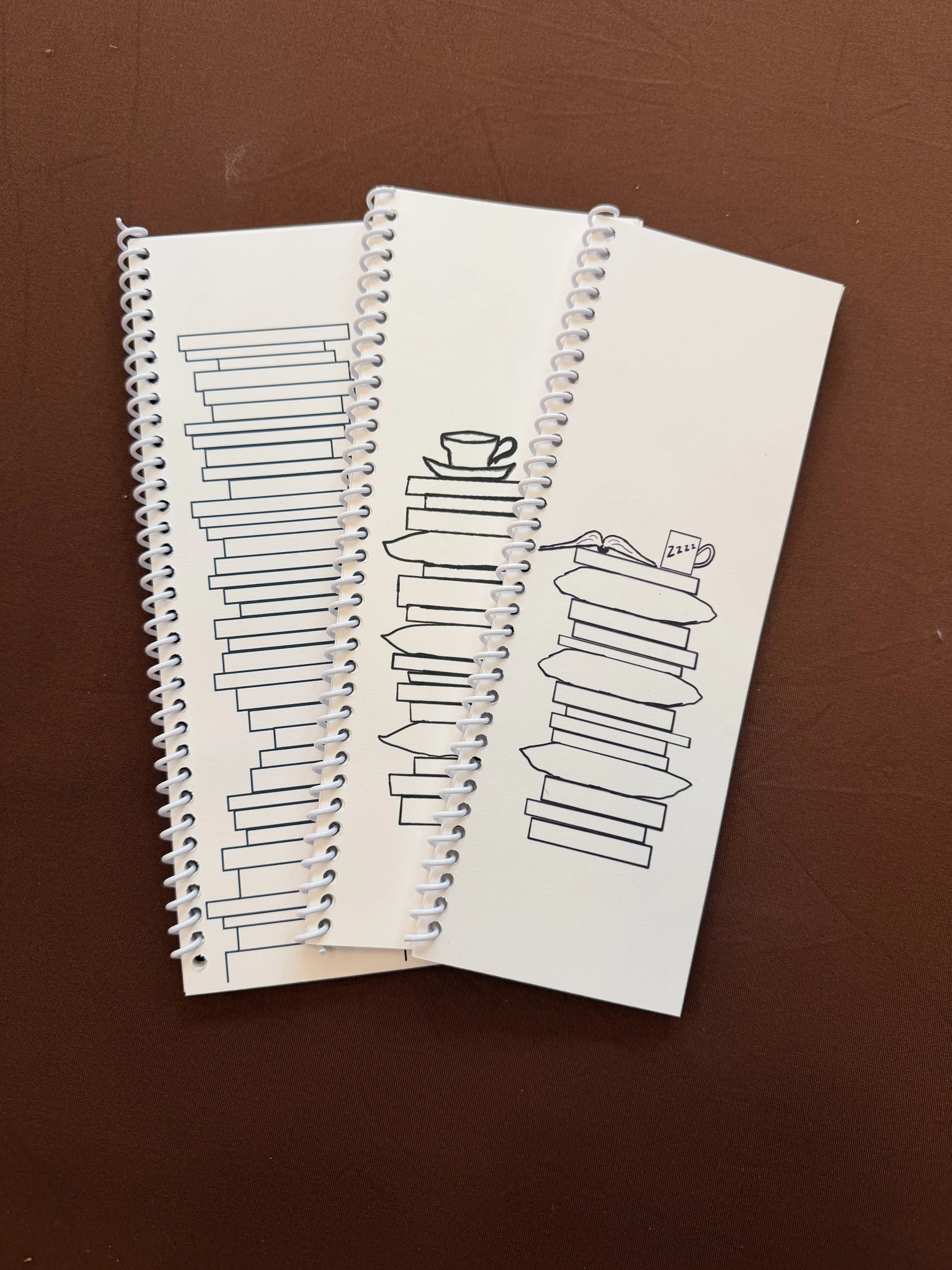 Three spiral-bound notebooks with minimalist black line drawings of stacked books and cups on the covers, placed on a brown surface.