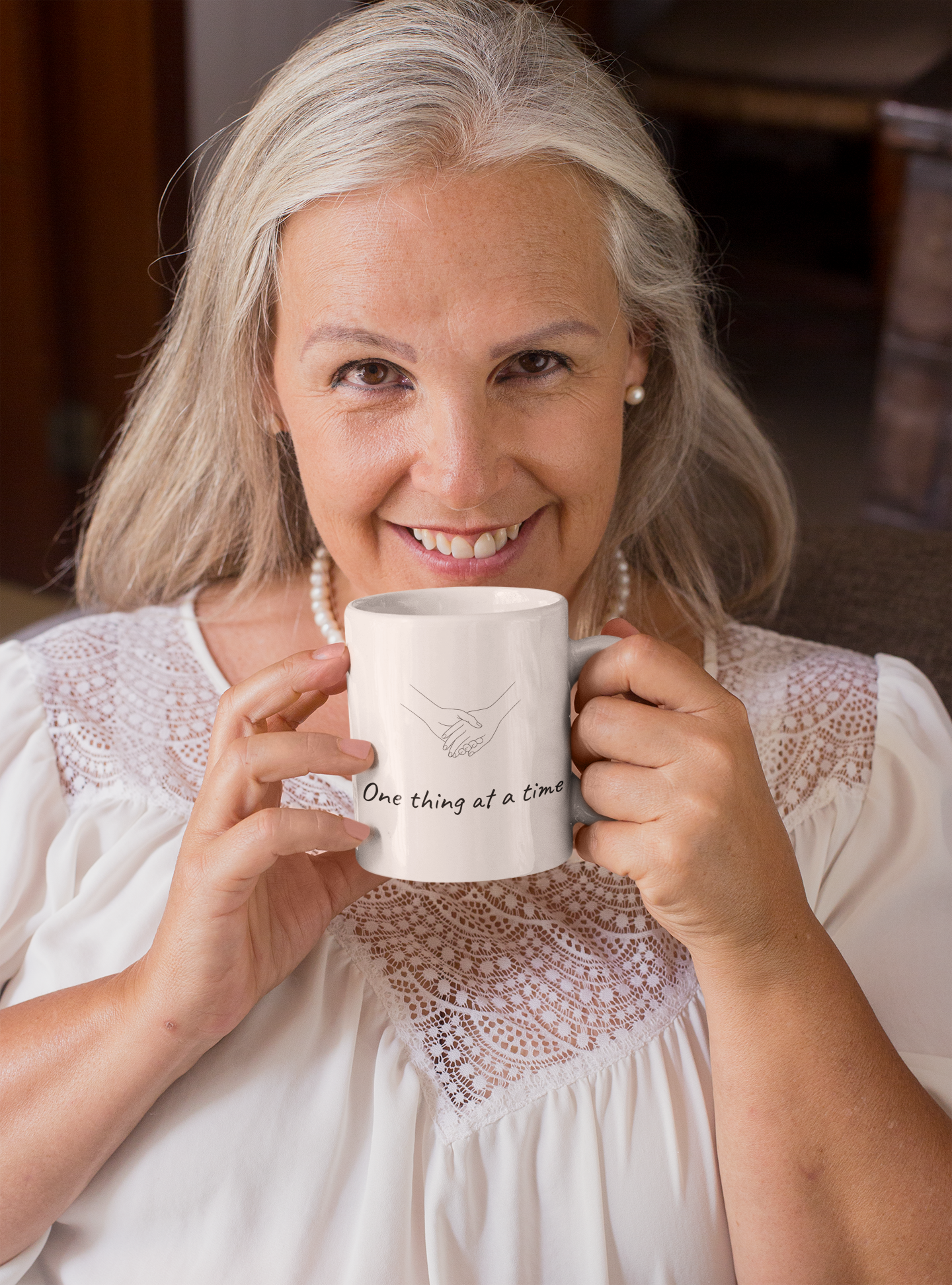 Smiling woman with gray hair holding a white mug with a handshake illustration and the phrase "One thing at a time" printed on it, sitting indoors.