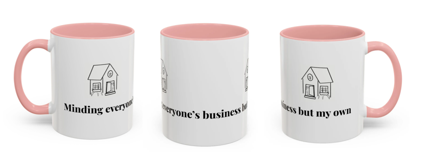 White ceramic mug with pink handle and interior, featuring a black line drawing of a house and black text splitting across three mugs that reads, "Minding everyone's business but my own".