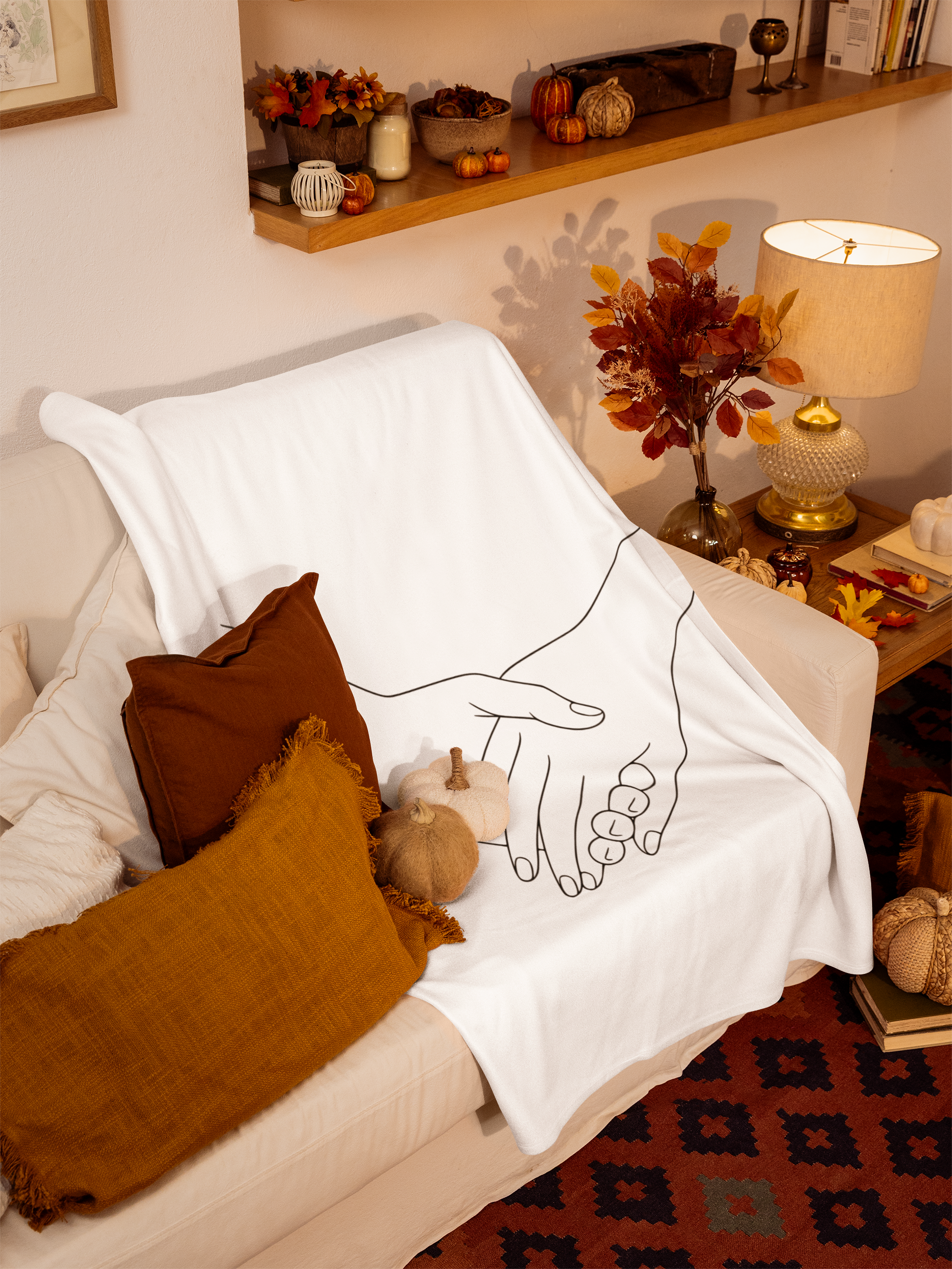 A cozy living room corner with a white sofa covered with a white cloth featuring a black line drawing of two hands clasped together. There are brown and mustard-yellow pillows on the sofa. To the side, there is a wooden side table with decorative pumpkins, a wicker basket, and fall-themed items. A table lamp with a cream lampshade and a textured glass base sits on the table, casting warm light. The background features a wooden shelf with pumpkins, gourds, flowers, candles, and books, with a patterned rug beneath.
