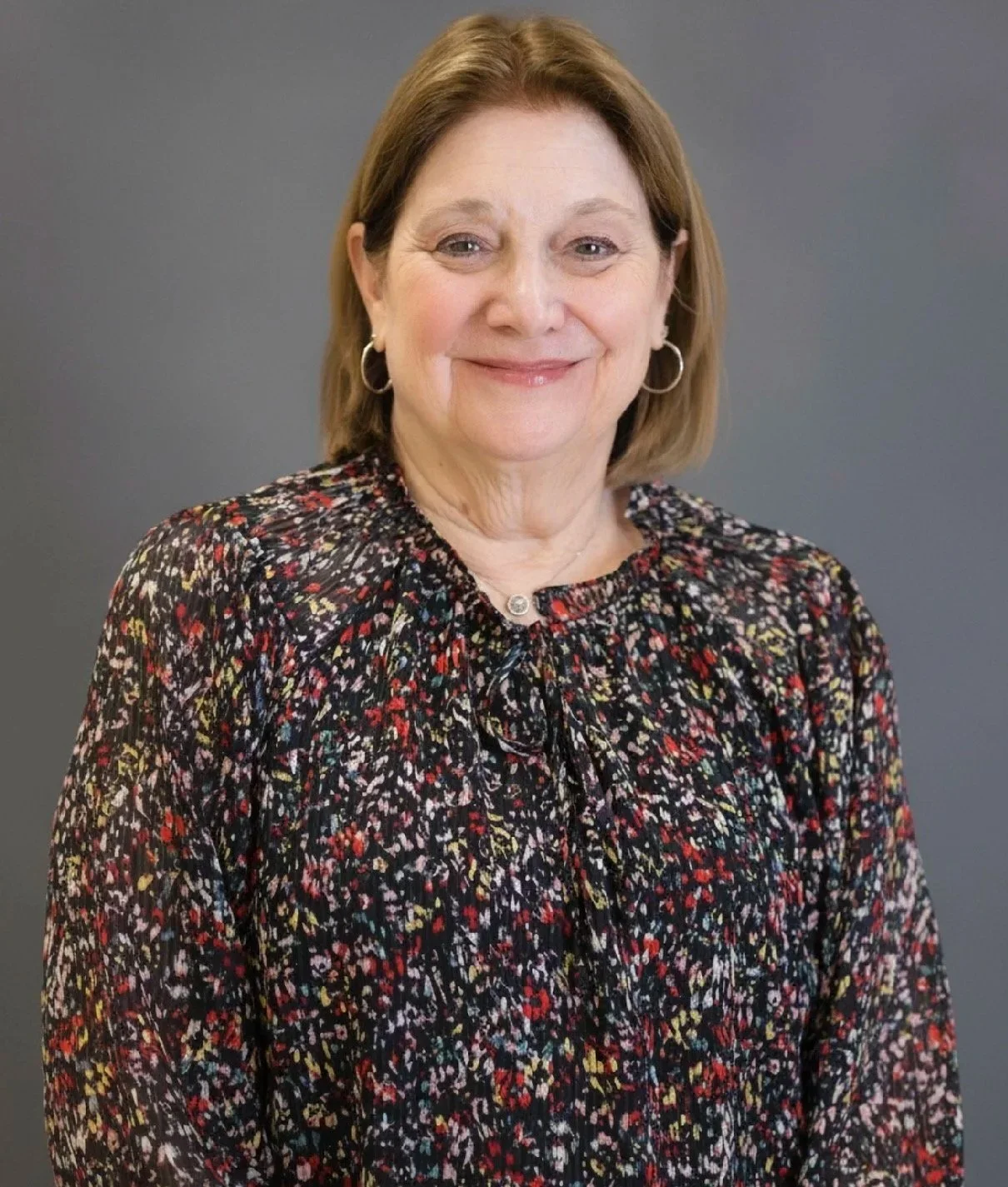 A middle-aged woman with short light brown hair wearing a multicolored floral blouse, silver hoop earrings, and a necklace, smiling at the camera against a dark gray background.