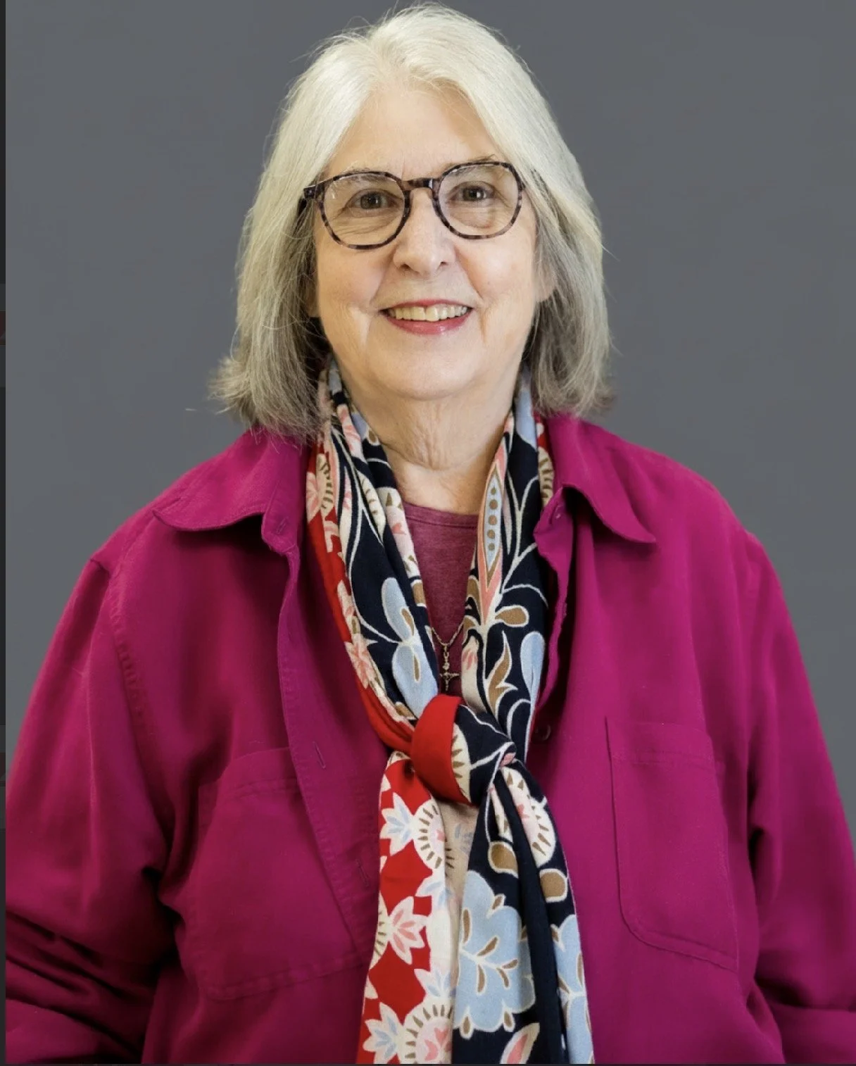A woman with gray hair wearing glasses, a pink shirt, and a floral scarf, standing against a gray background.