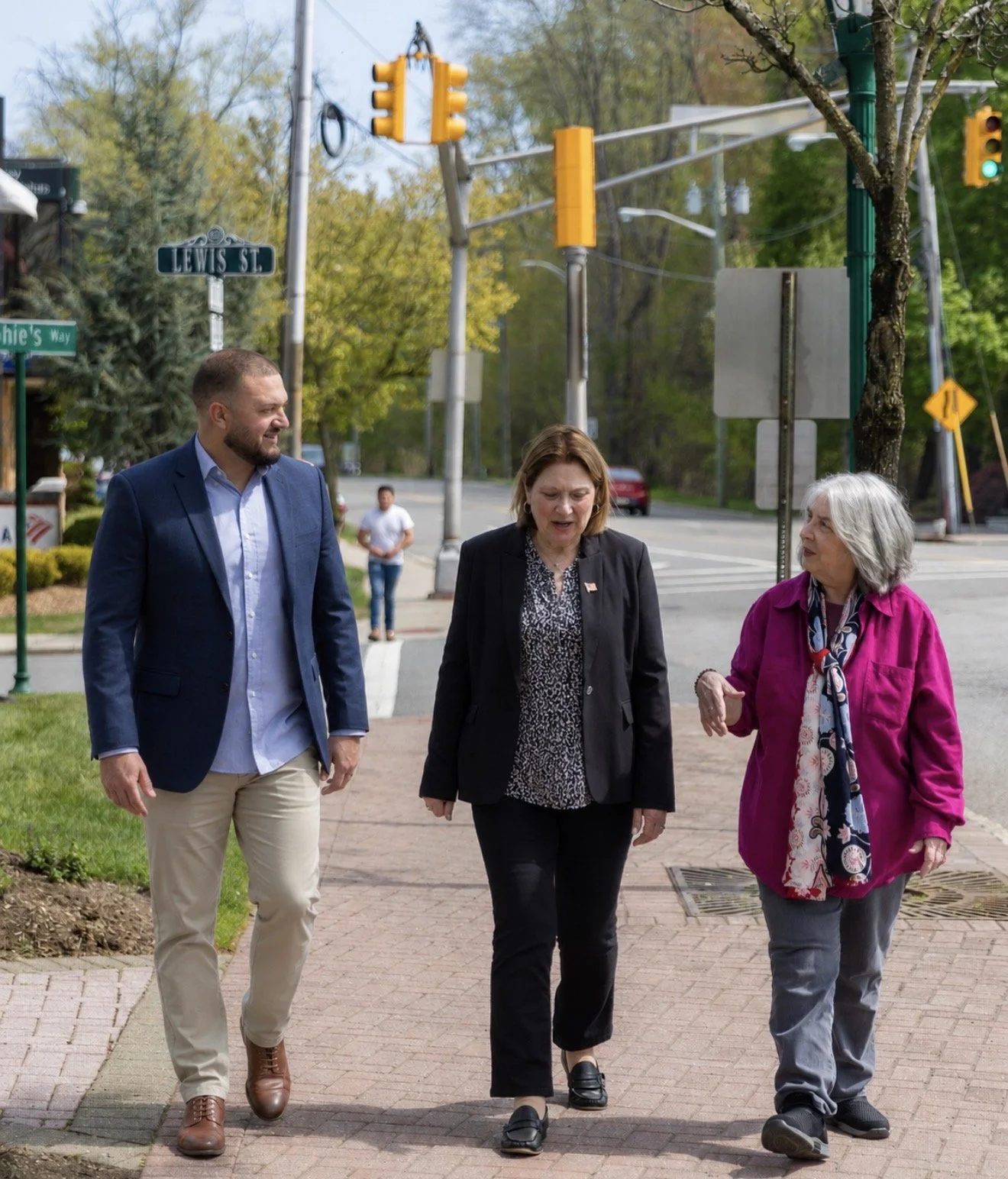 Three women and one man walking on a sidewalk in a city area during daytime, engaging in conversation. The women are wearing a pink jacket, black blazer, and a black blazer with patterned blouse, respectively. The man is dressed in a blue blazer, light blue shirt, and beige pants.