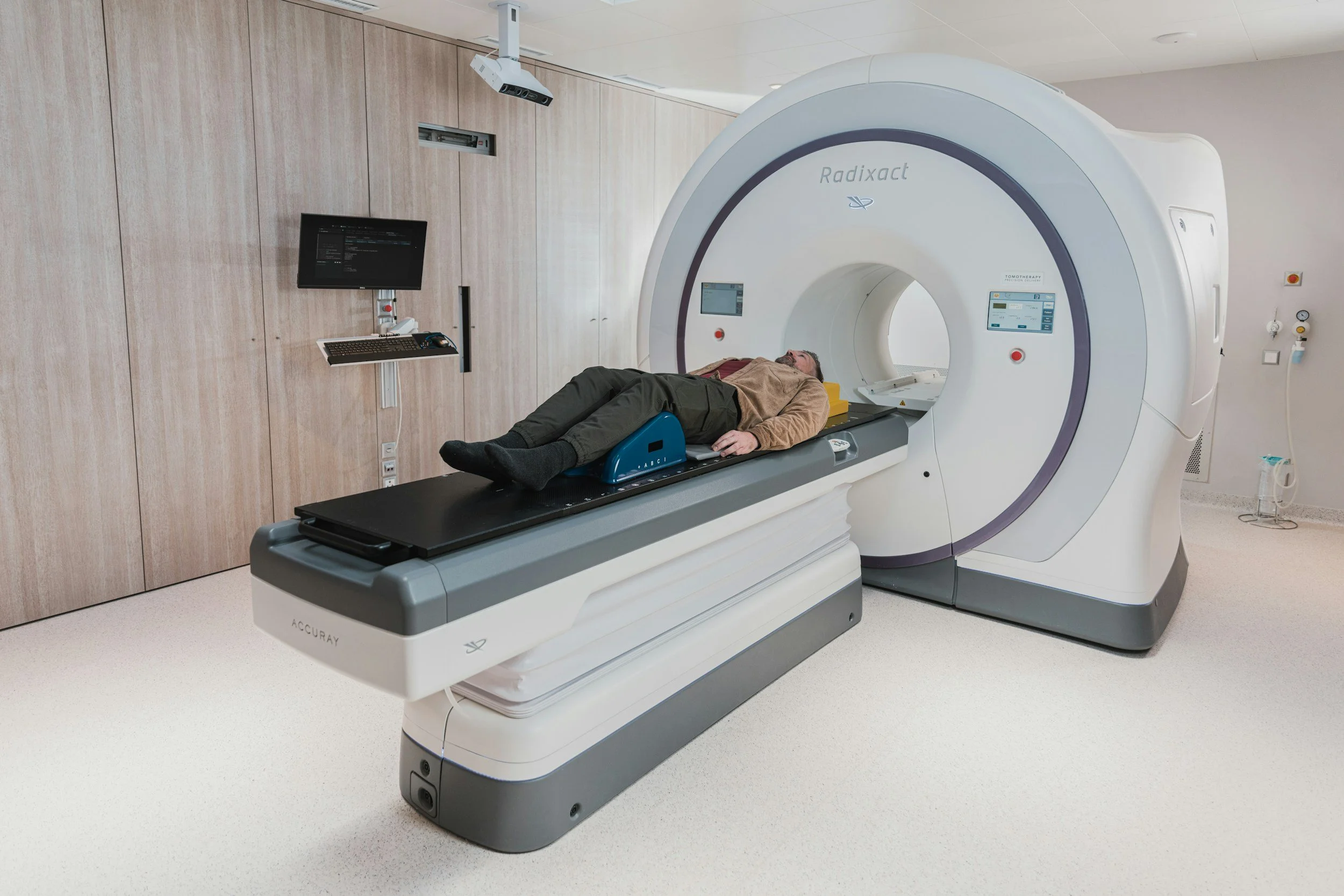 A person lying on a medical table inside a large radiation therapy machine labeled 'Radixact' in a hospital room with wooden wall panels and medical equipment nearby.