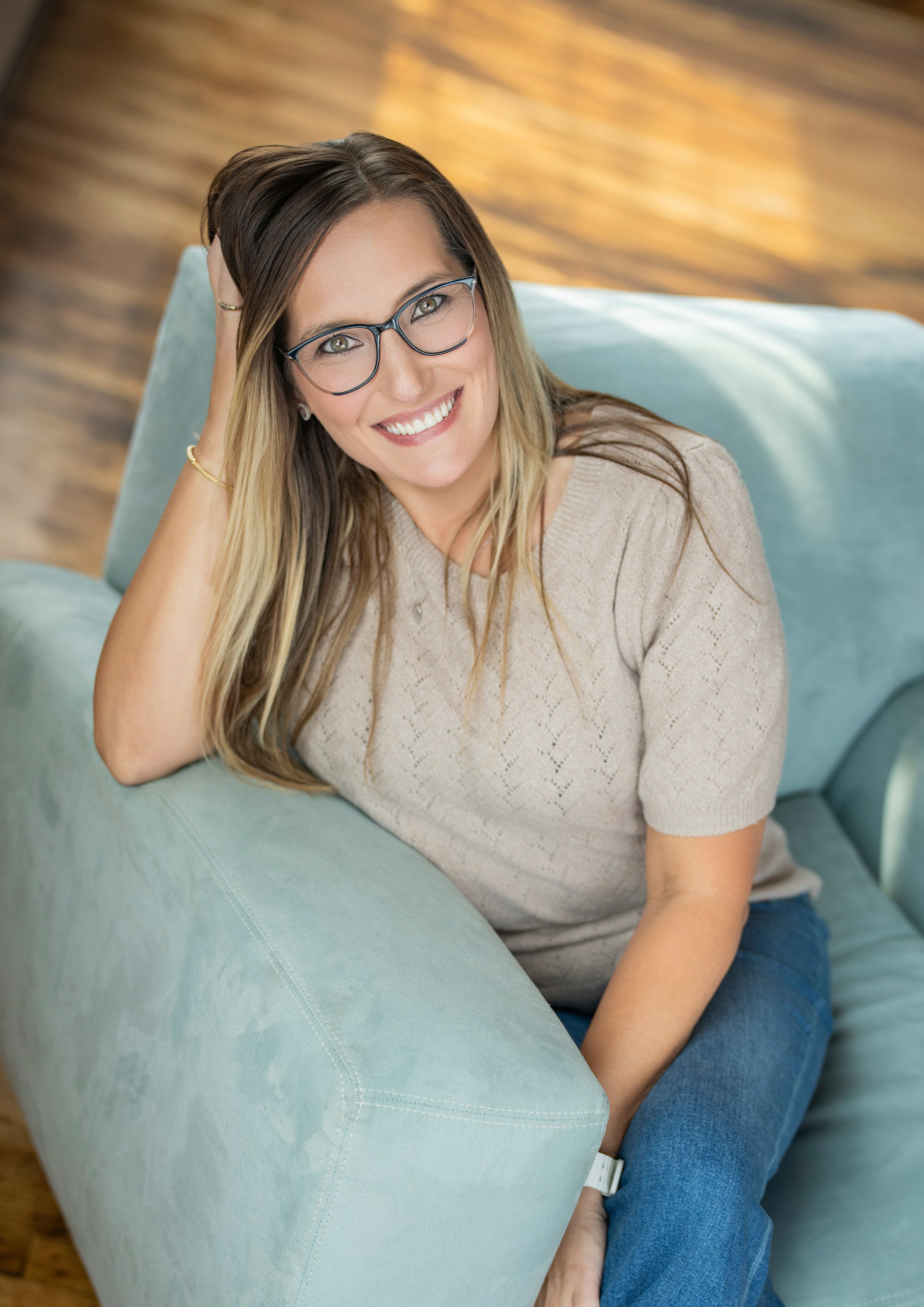 A woman with glasses and long hair sitting on a light blue couch, smiling at the camera, resting her head on her hand, with a wooden floor in the background.