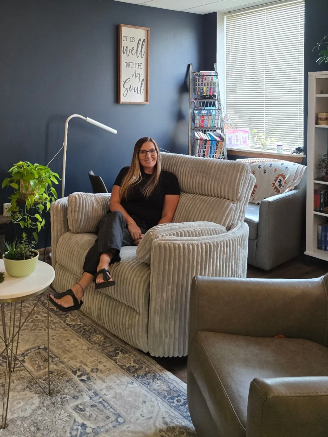 A woman sitting on a striped beige sofa in a cozy living room with a dark blue wall, a framed quote, potted plants, a brown chair, and a window with blinds.