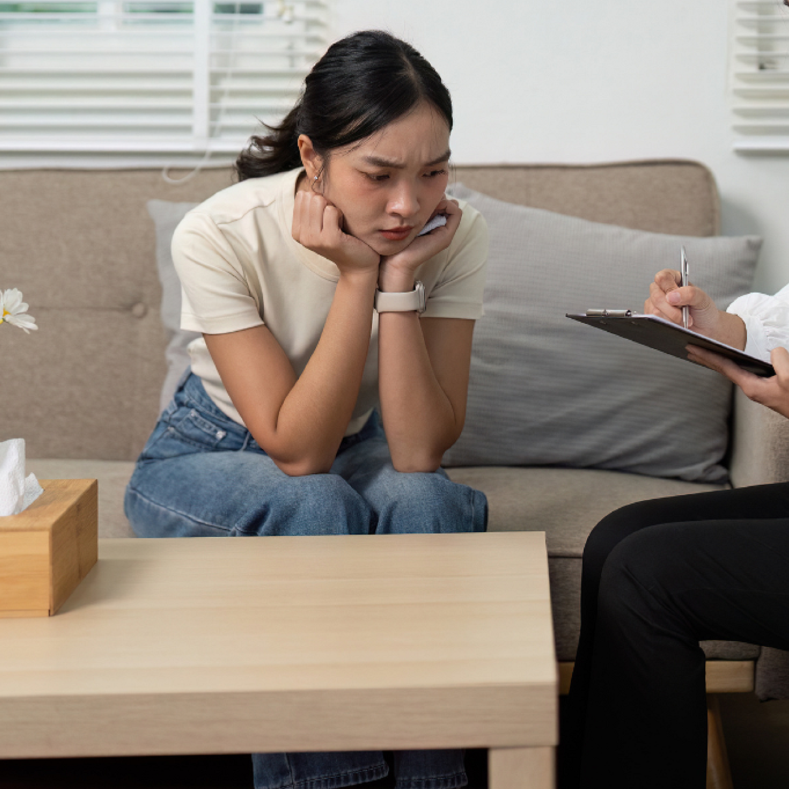 A young woman sitting on a beige couch with her elbows resting on her knees and hands on her cheeks, looking distressed. A person next to her is taking notes on a clipboard with a pen. The room has white blinds in the background and a wooden table with a tissue box and a flower.