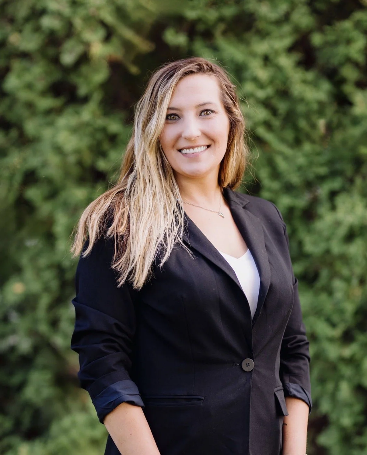 A smiling woman with long, wavy blonde hair wearing a black blazer and white top, standing outdoors in front of green foliage.