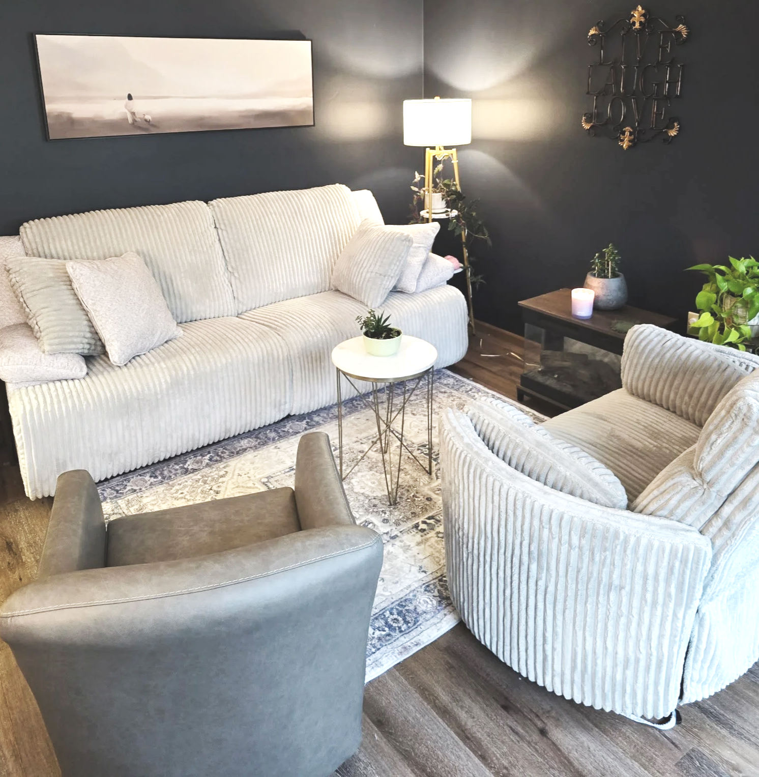 Living room with two off-white, ribbed fabric sofas, a gray armchair, and a small round white side table with a potted plant. Dark walls with a large monochromatic landscape painting, a wall sign reading 'Live, Laugh, Love,' a floor lamp, and various potted plants on a side table and coffee table. A patterned area rug is underneath the furniture, and the room has wooden flooring.