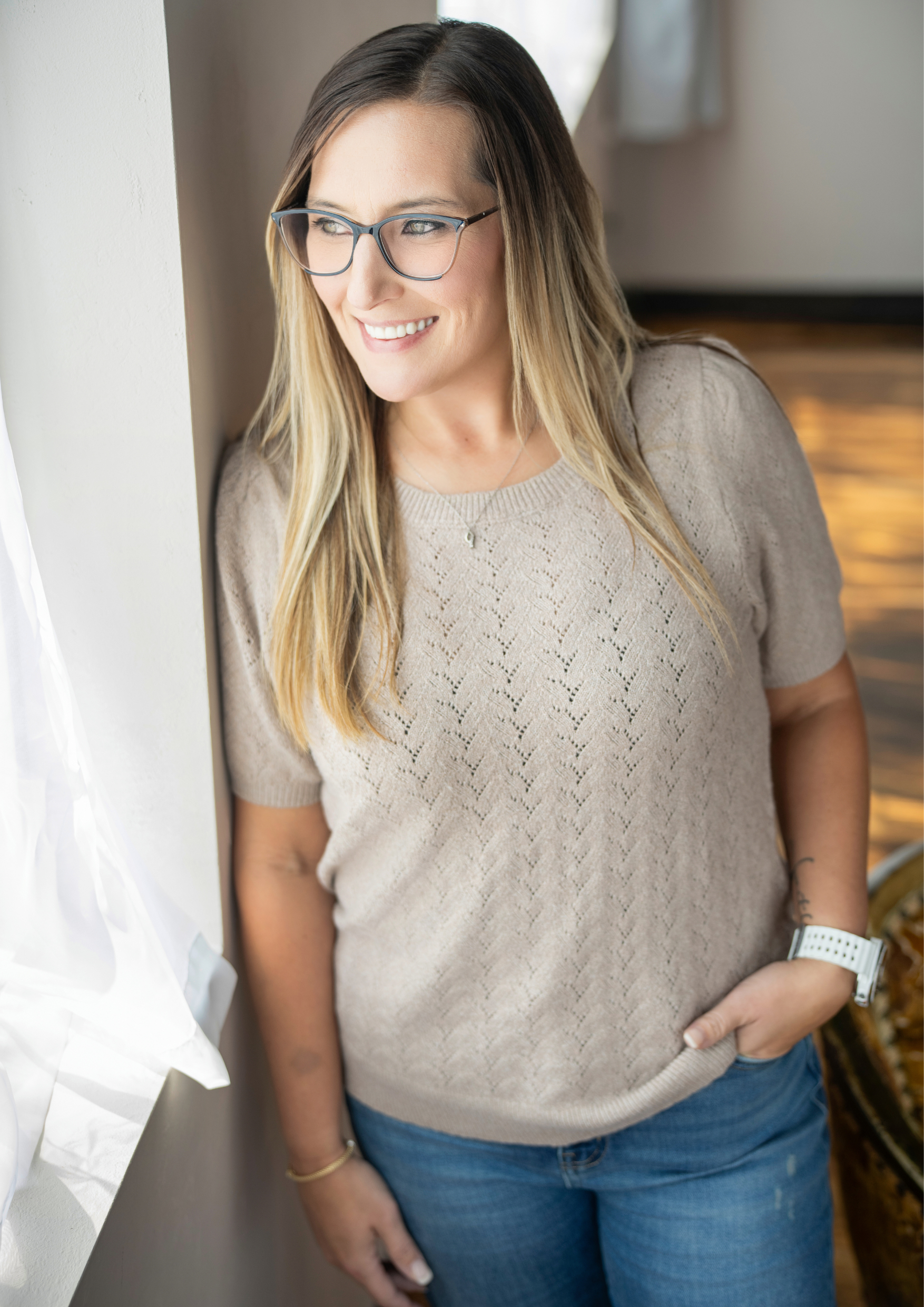 A woman with long blonde hair wearing glasses and a beige sweater, standing indoors, smiling and looking to the side.