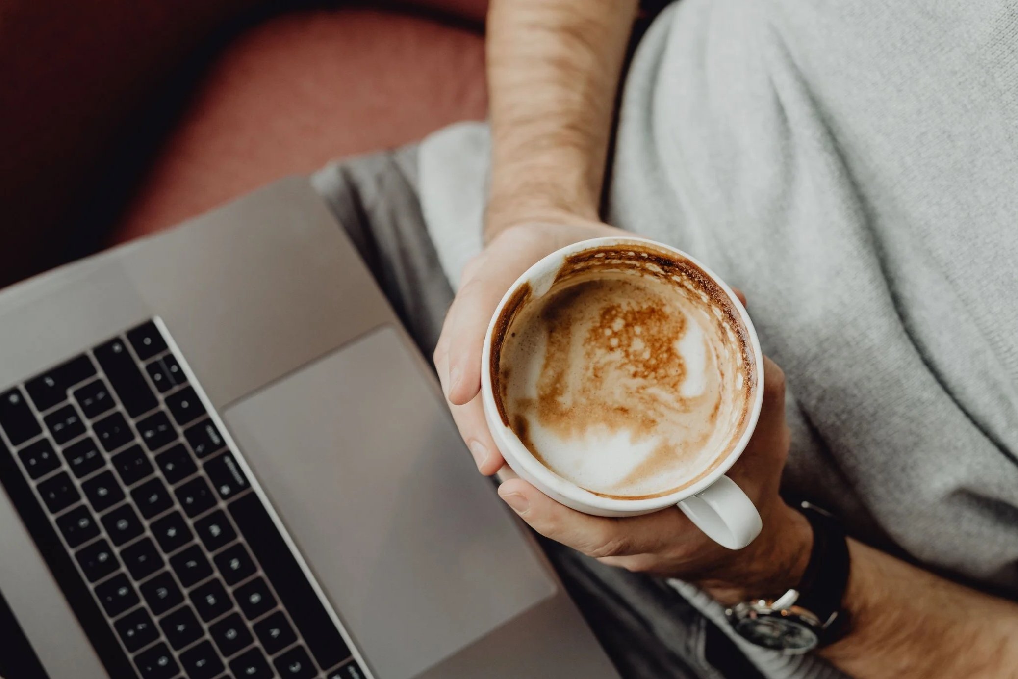 A person holding a partially empty cup of coffee or latte with foam on top, with a laptop keyboard visible in the background.