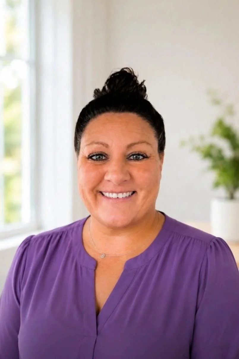 A woman with dark hair tied up in a bun, smiling, wearing a purple top, indoors near a window with a blurred green plant in the background.