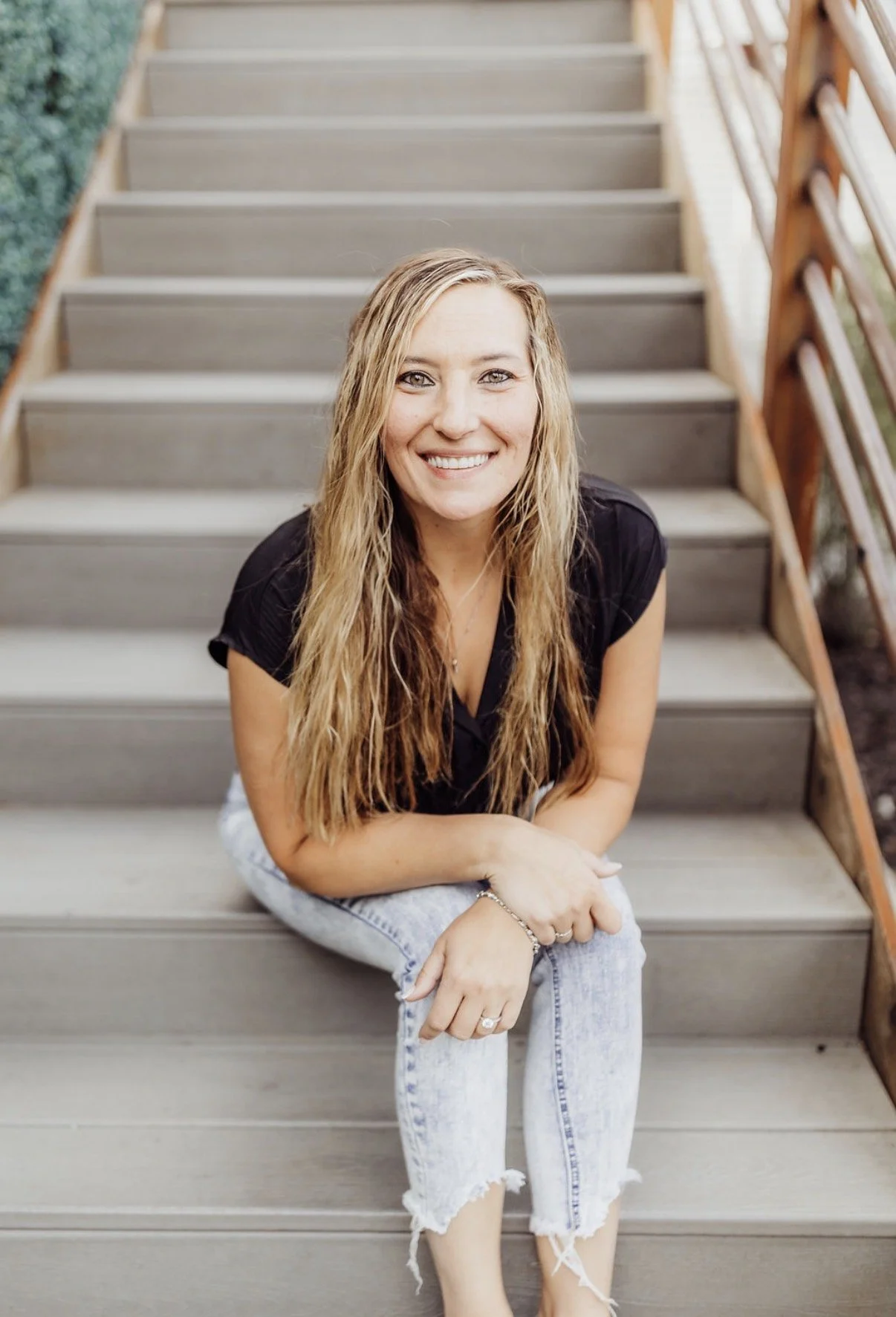 A woman with long, wavy blonde hair smiling and sitting on outdoor wooden stairs, wearing a black top and ripped light-wash jeans.