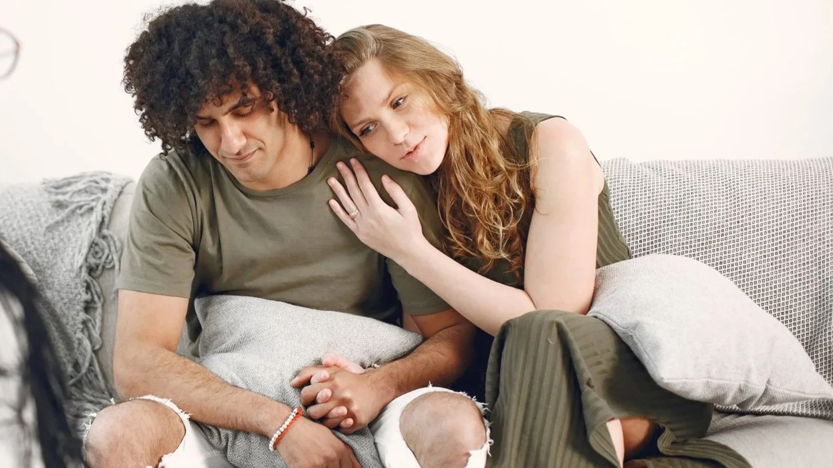 A couple sitting closely on a sofa watching something, with the woman resting her head on the man's shoulder and her hand on his chest.