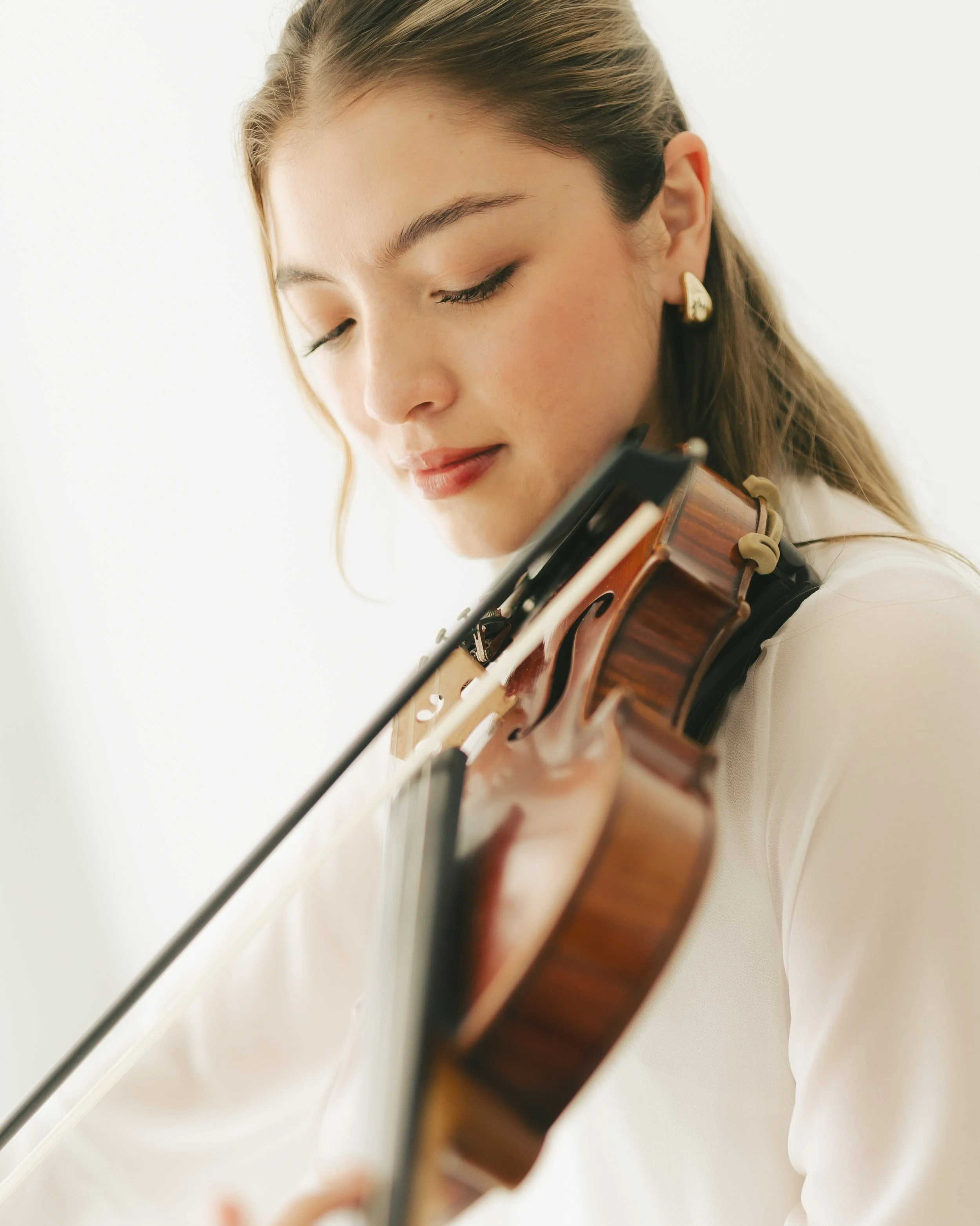 Close-up of a young woman with light brown hair playing a violin, wearing gold earrings, against a plain light background.
