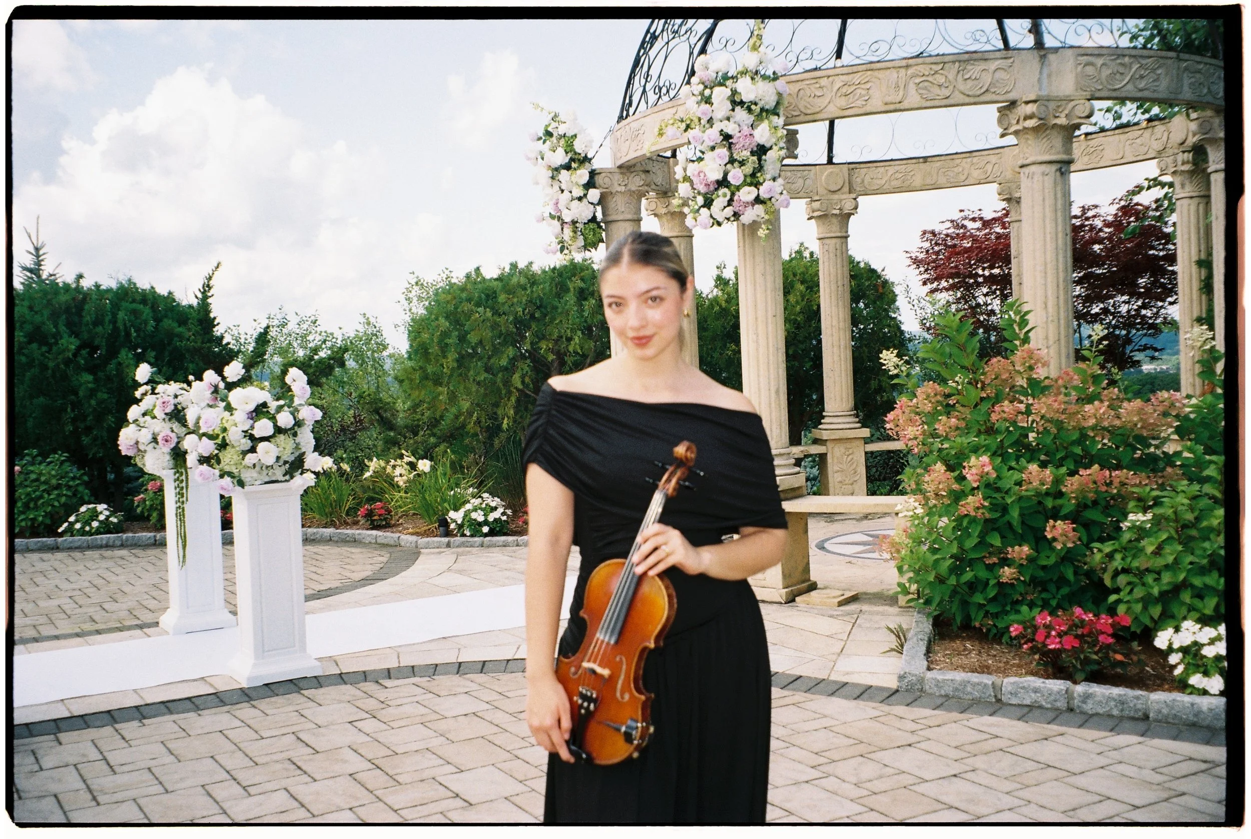 A woman in a black off-shoulder dress holding a violin, standing outdoors in front of a decorative stone and metal structure with floral arrangements at a daytime event.