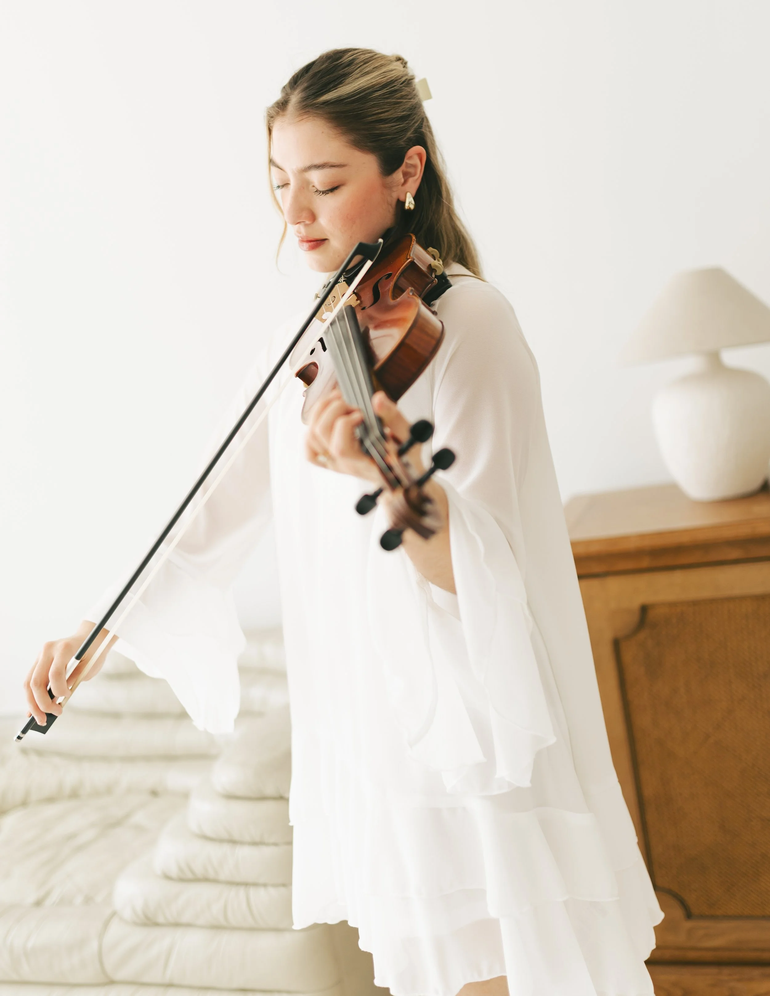 Young woman playing the violin indoors, wearing a white dress with a white background and wooden furniture.