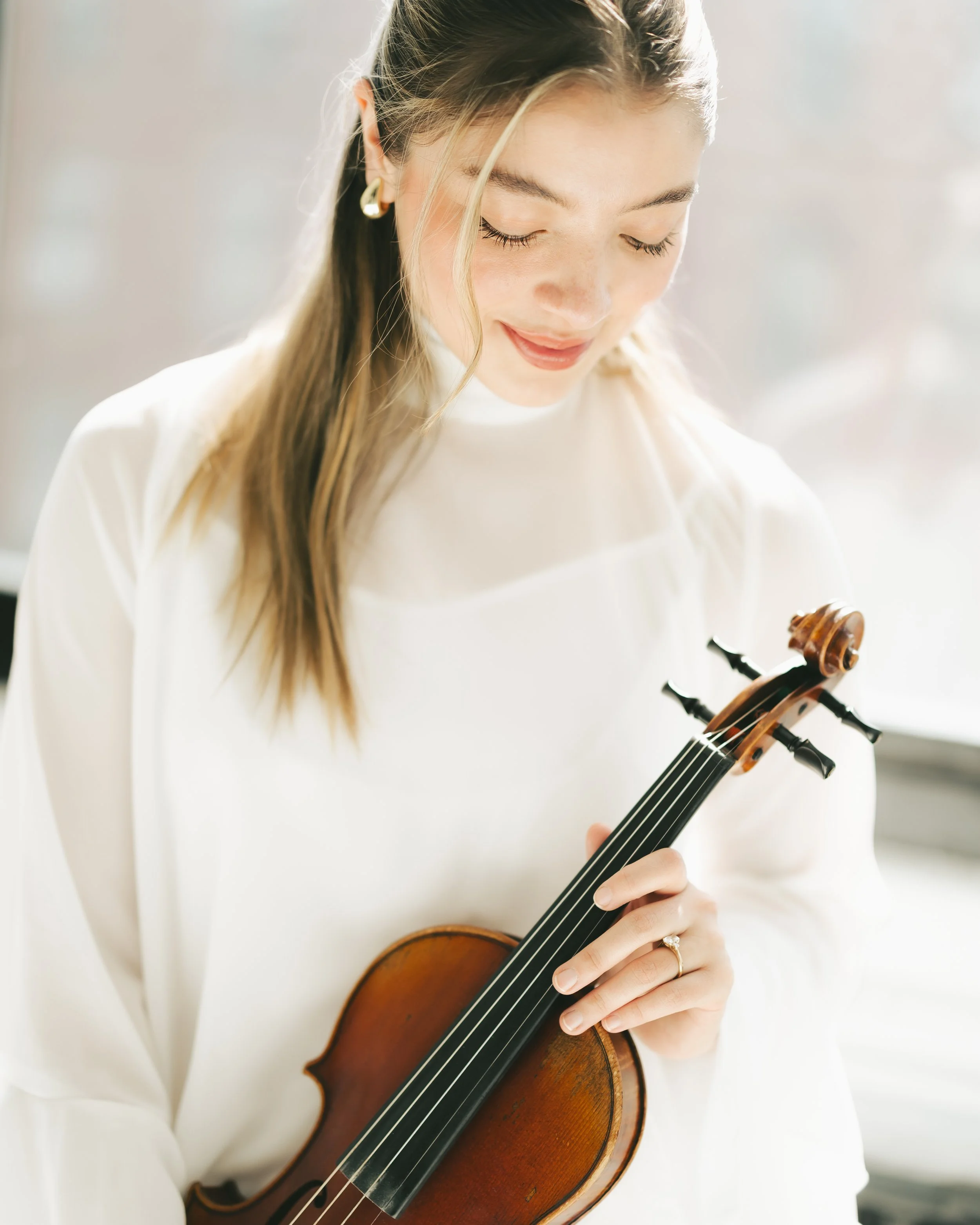 A woman with long blonde hair playing a violin, wearing a white top and gold earrings, with a soft smile on her face.