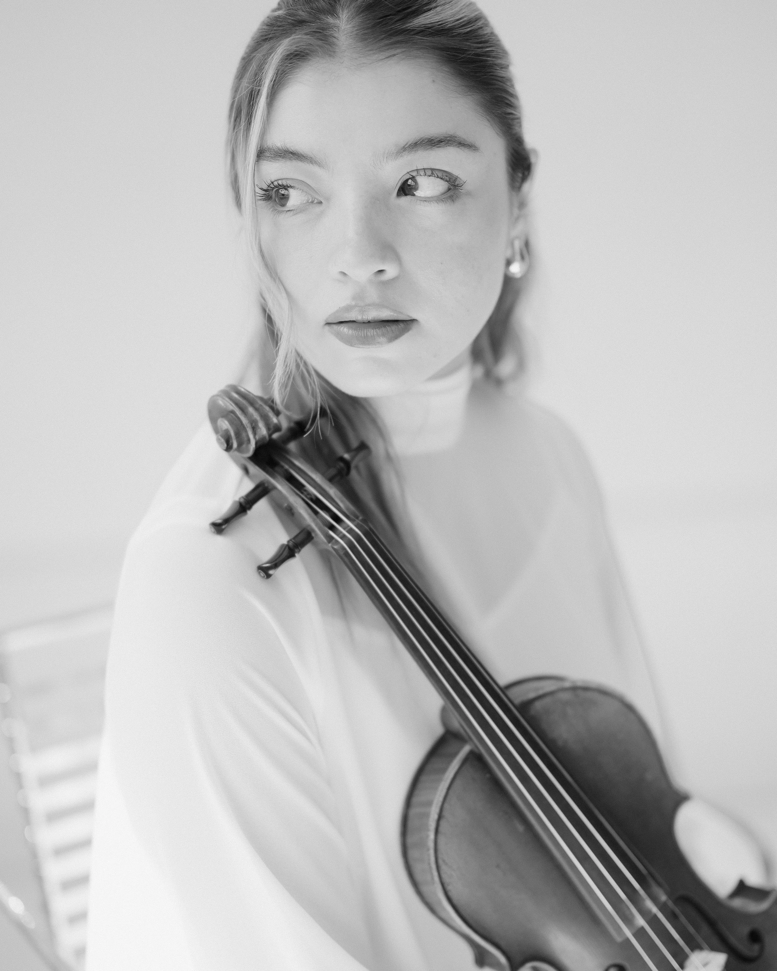 Close-up of a woman holding a violin and looking to the side, with a neutral background.