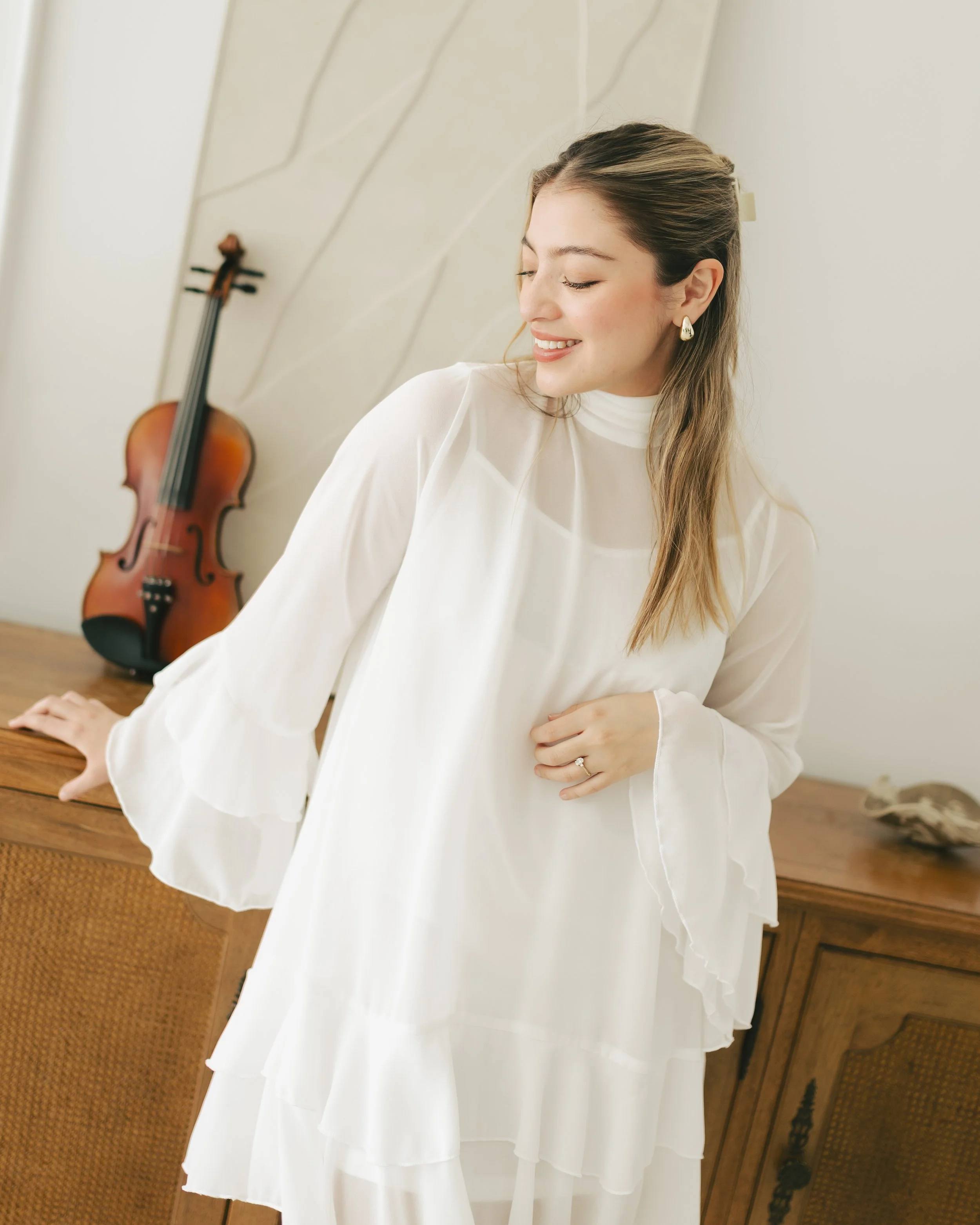A woman with long light brown hair styled in a half-updo, wearing a white high-neck blouse and teardrop-shaped earrings, smiling with her eyes closed, standing in front of a cream-colored wall with diagonal paneling, and a wooden dresser with a violi