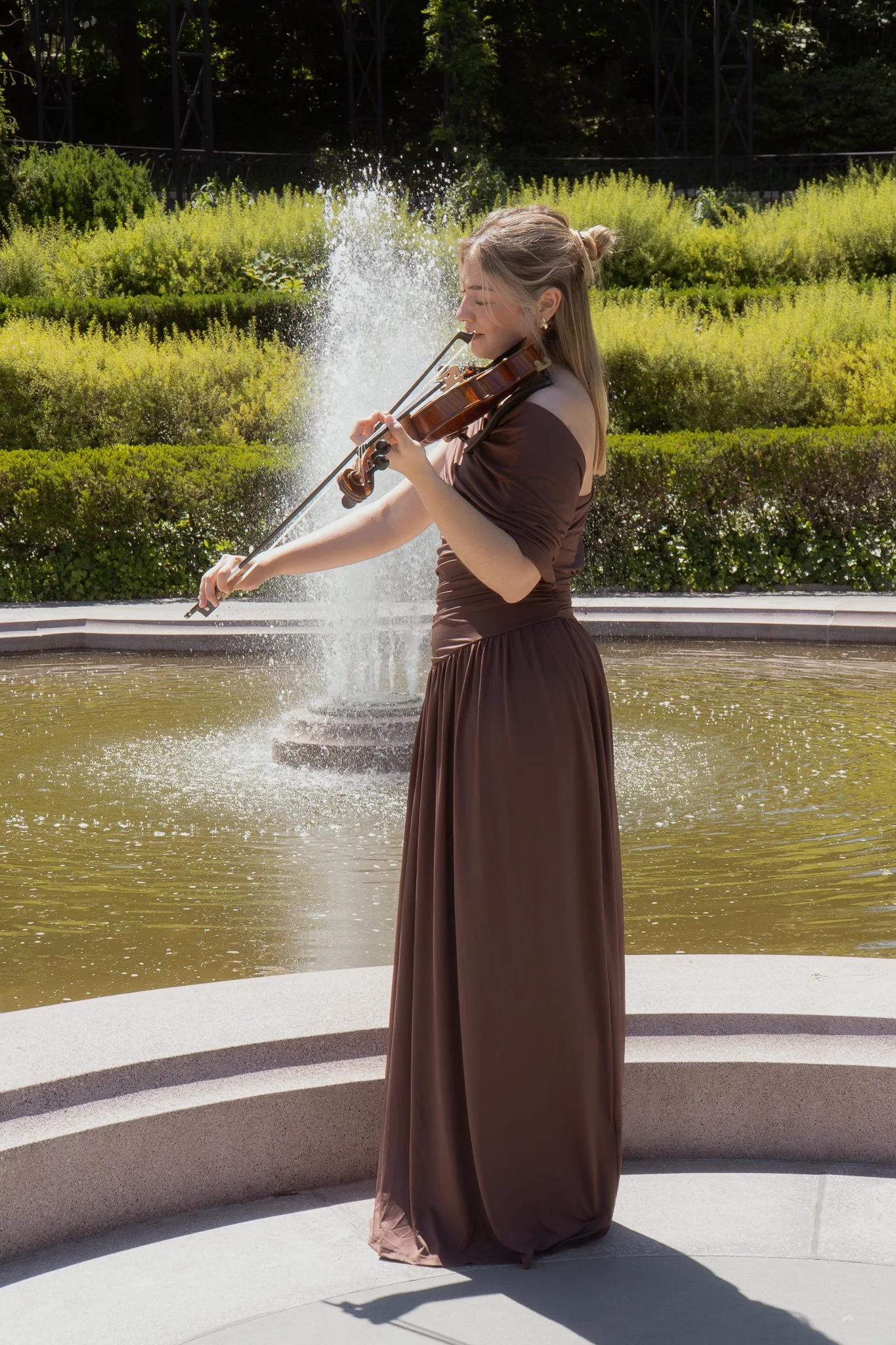 A woman in a brown dress playing the violin outdoors near a fountain with greenery in the background.