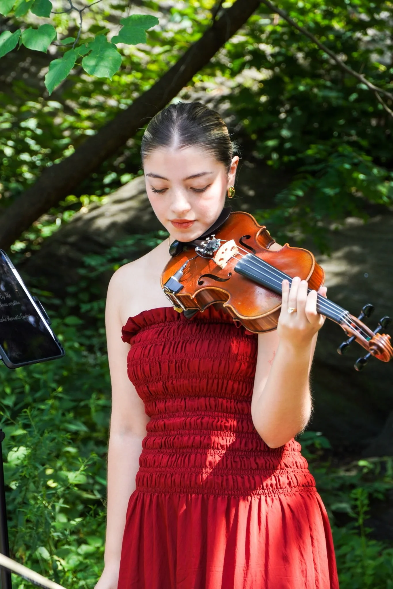 A young woman wearing a red strapless dress playing a violin outdoors in a green, wooded area.