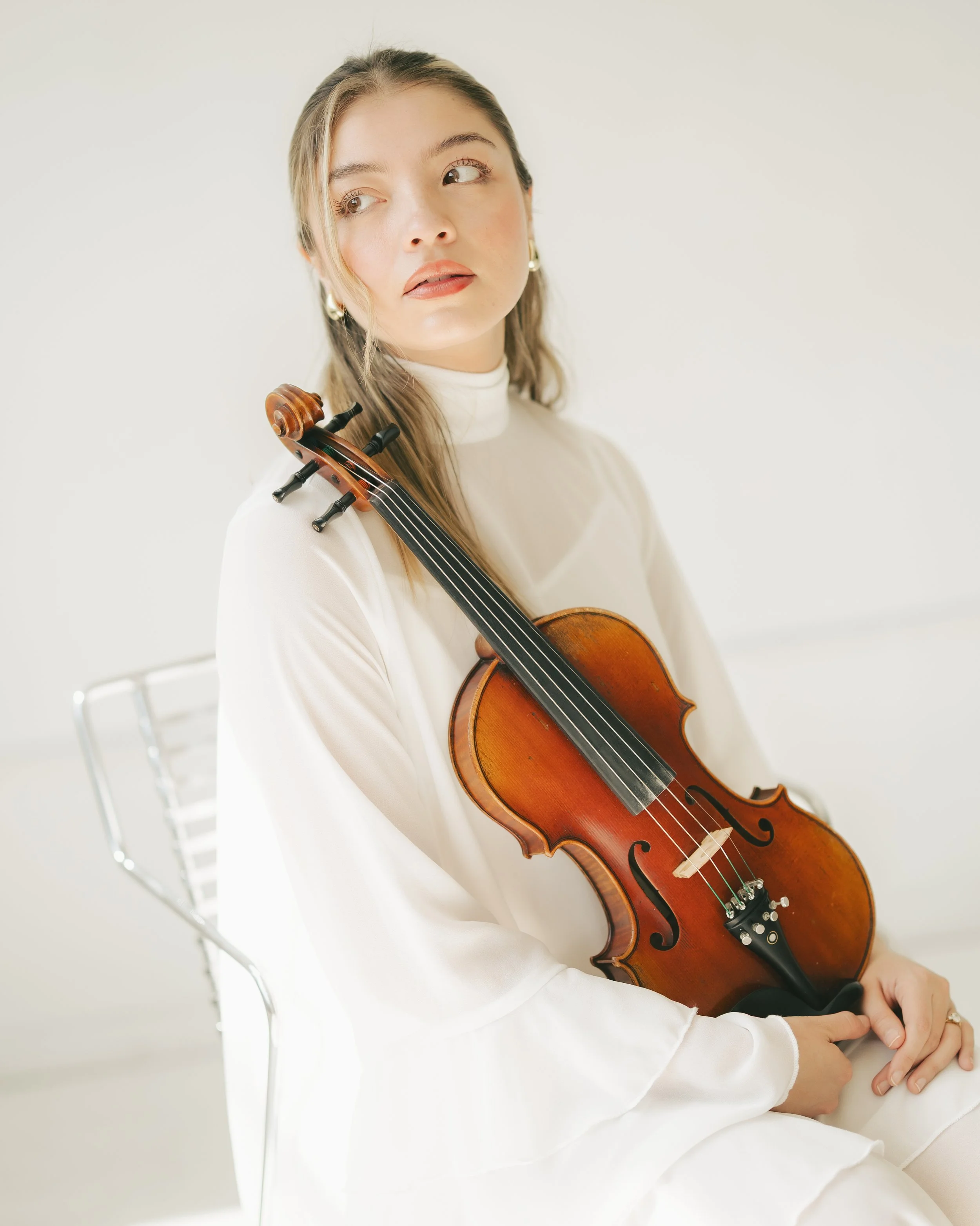 A woman with long, light brown hair wearing a white turtleneck, sitting on a white chair and holding a violin while looking to her left in a bright, minimalistic room.