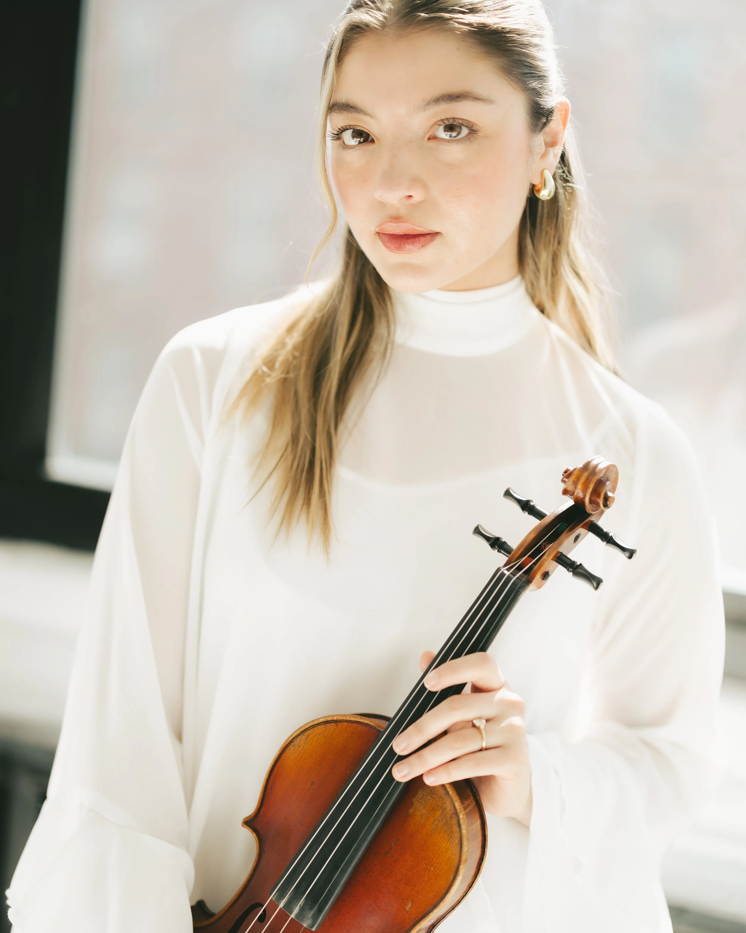 A young woman with light brown hair wearing a white top, holding a violin in her left hand, and looking attentively at something off-camera.
