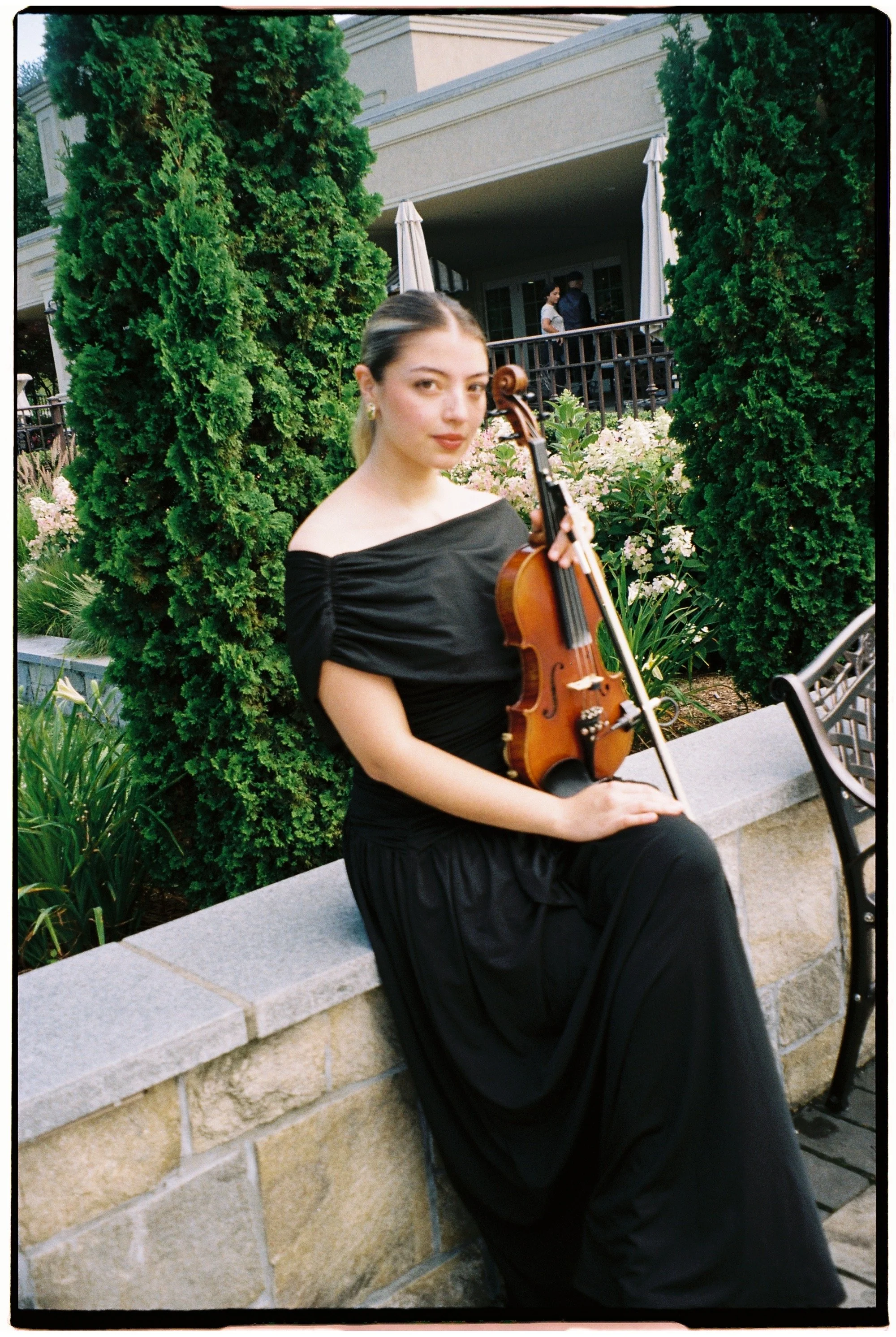A young woman sitting on a stone ledge outdoors, holding a violin, with tall green trees and a building with a balcony in the background.